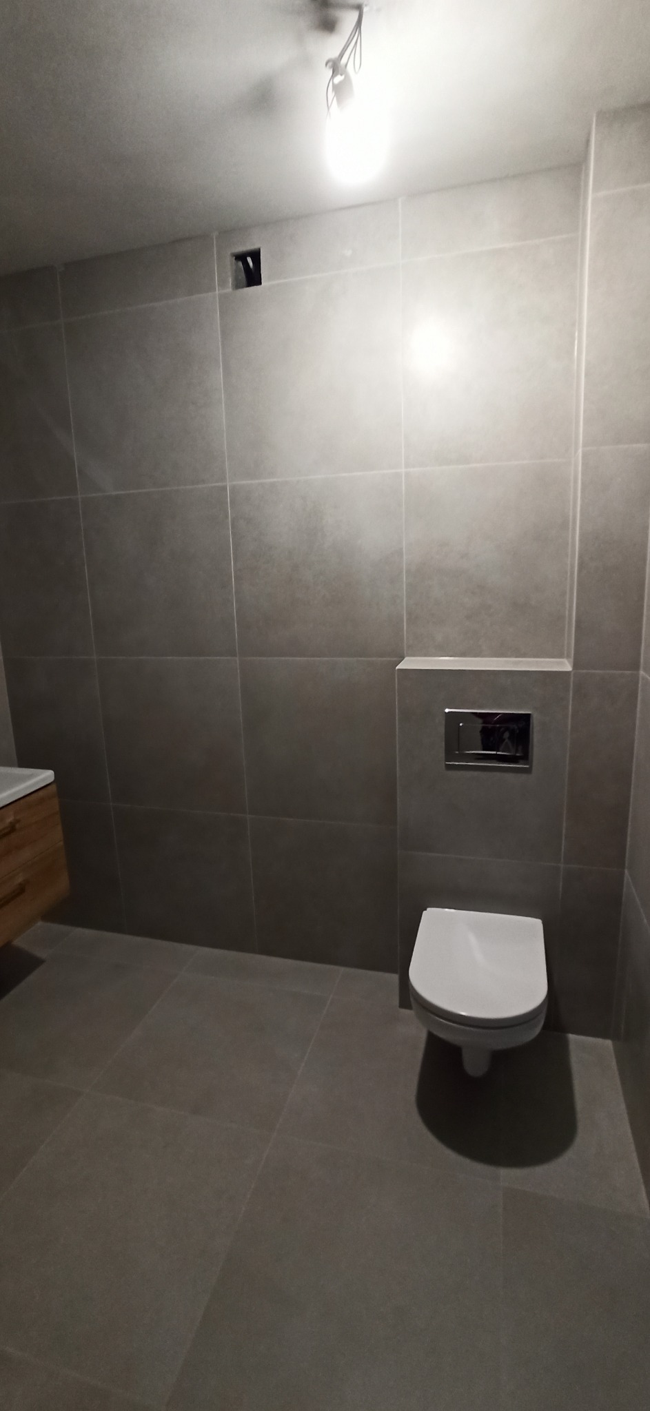 Nowoczesna łazienka z szarymi, kwadratowymi płytkami na ścianach i podłodze; wisząca toaleta z chromowanym przyciskiem spłukującym; jasne oświetlenie sufitowe; częściowo widoczna drewniana szafka...