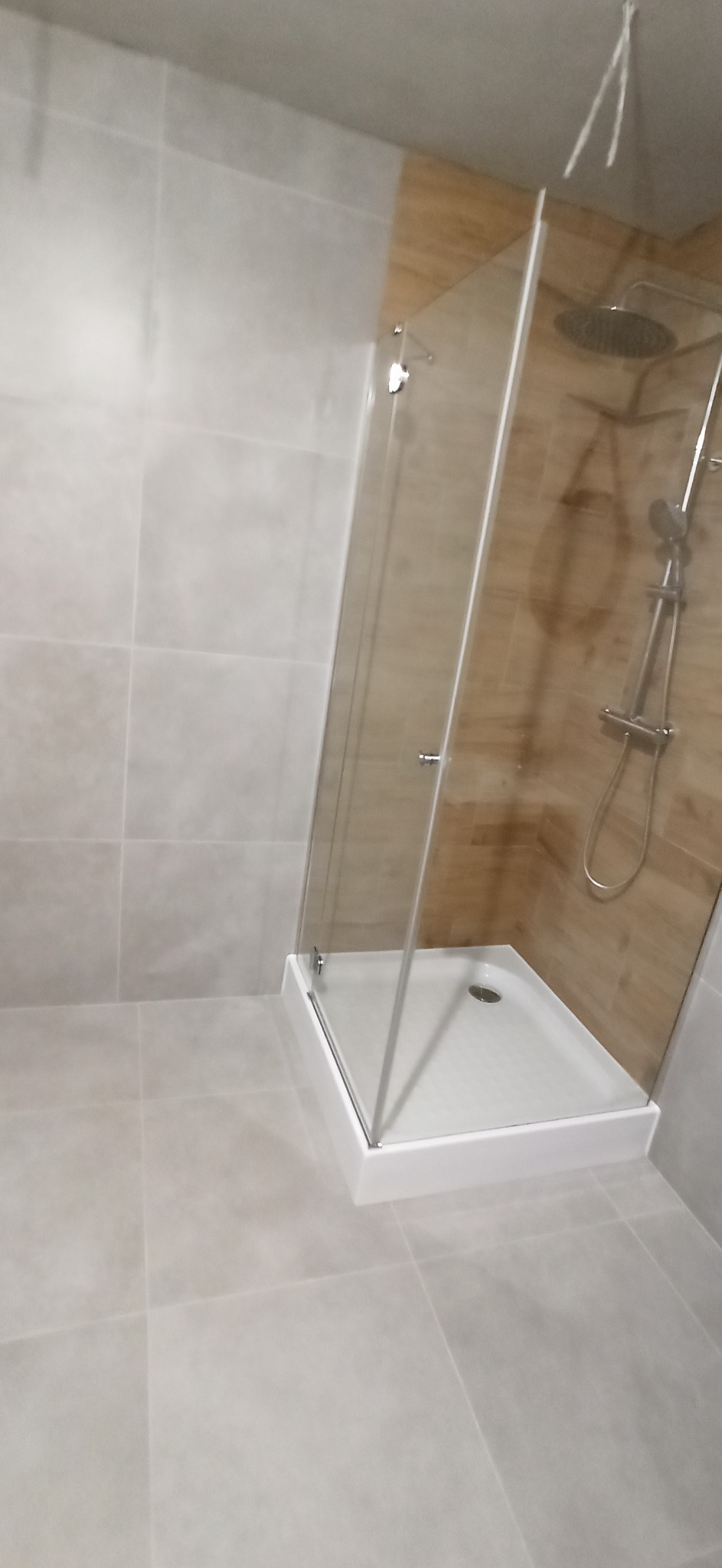 Nowoczesna kabina prysznicowa z deszczownicą, obudowana płytkami imitującymi drewno, widok z boku na szare płytki podłogowe i ścienne.