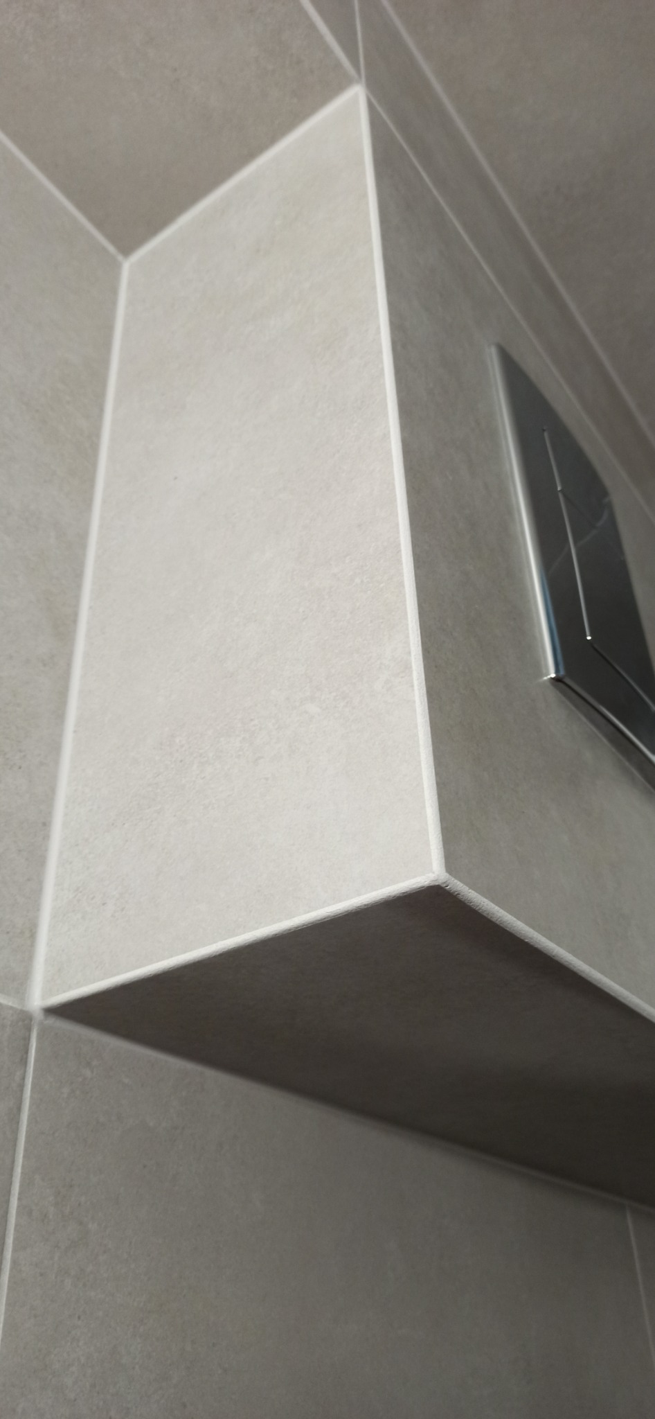 Szare, matowe płytki ceramiczne w narożniku łazienki z widocznym przyciskiem spłuczki w zabudowie podtynkowej