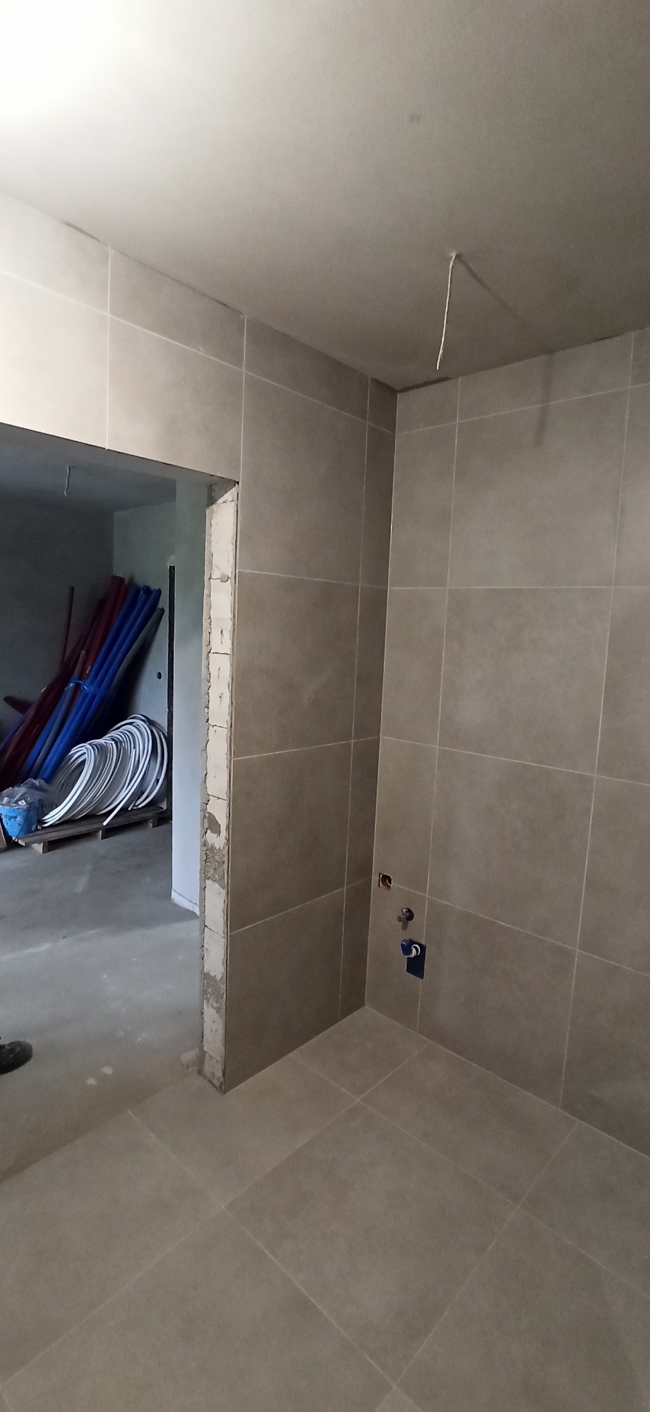 Świeżo wykafelkowana łazienka w odcieniach szarości, widoczne przyłącza wodne, w tle rury i materiały budowlane w otworze drzwiowym.