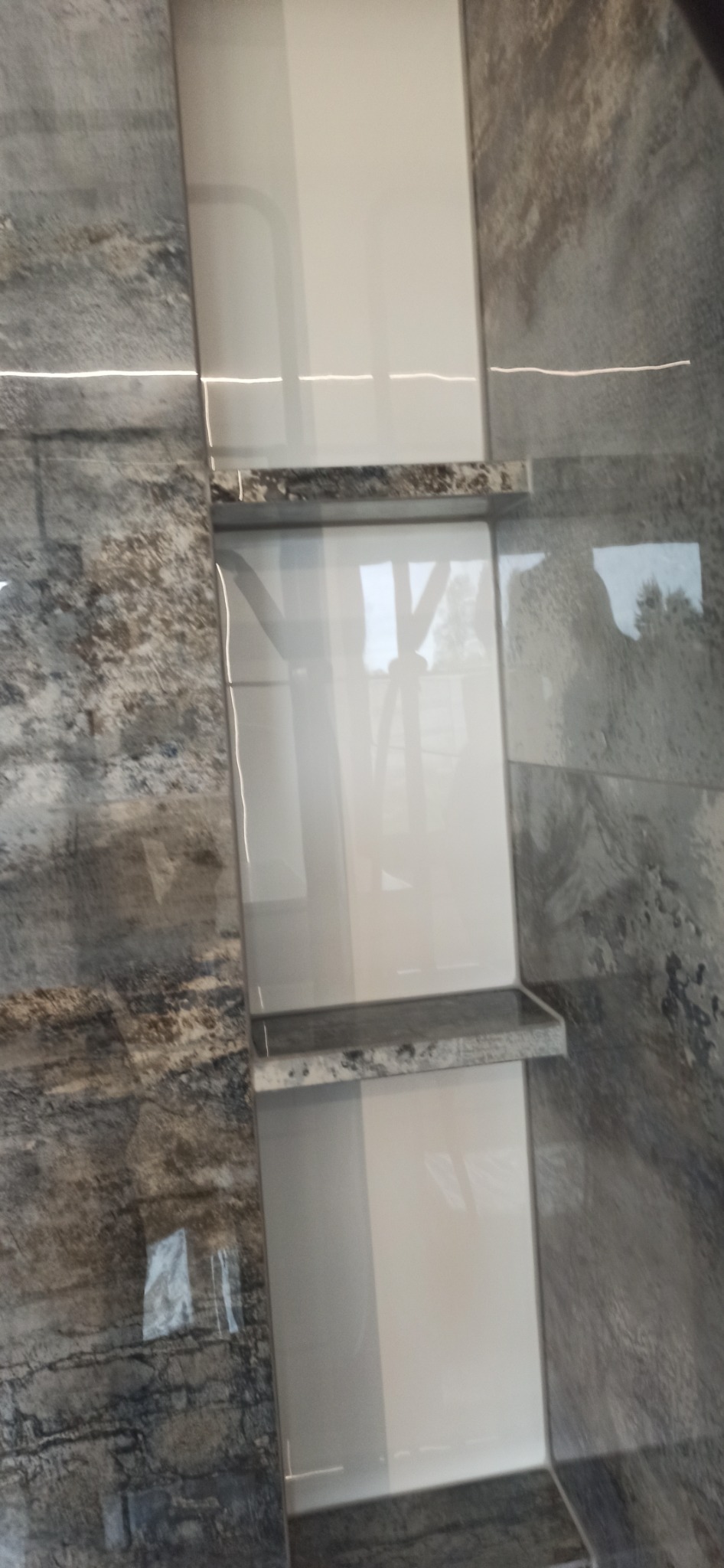 Wnęka w łazience wyłożona białymi płytkami, z dwiema półkami z szarego kamienia, otoczona płytkami imitującymi kamień w odcieniach szarości i brązu.