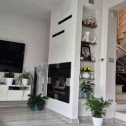 Salon z nowoczesnym kominkiem w zabudowie, ozdobnymi półkami, białymi ścianami i szarą podłogą imitującą drewno, doświetlony naturalnym światłem.