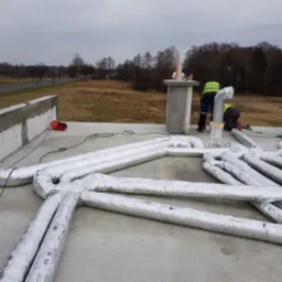 Montaż izolowanych przewodów wentylacyjnych na dachu budynku w trakcie budowy; widoczni dwaj pracownicy w kamizelkach odblaskowych, ceglany komin i narzędzia.
