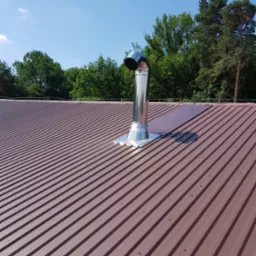 Brązowy dach z blachy trapezowej z zamontowanym, błyszczącym, metalowym kominem wentylacyjnym na tle zielonych drzew i błękitnego nieba.