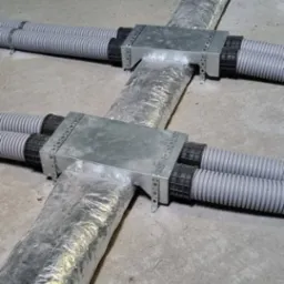 Krzyżowe połączenie przewodów wentylacyjnych z metalowymi elementami mocującymi, widoczne rury karbowane i izolacja folią aluminiową na surowym betonowym podłożu.