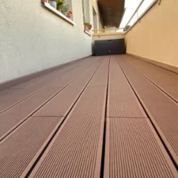 Nowoczesny balkon wyłożony panelami kompozytowymi w odcieniach brązu, z widocznym pojemnikiem na poduszki i kwiatami w doniczkach na parapecie.