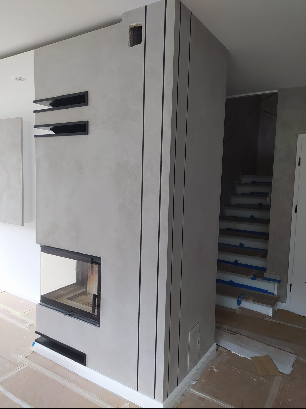 Nowoczesny kominek w obudowie z szarego betonu architektonicznego, z czarnymi, pionowymi paskami dekoracyjnymi i półkami wnękowymi, widoczne schody zabezpieczone taśmą malarską.