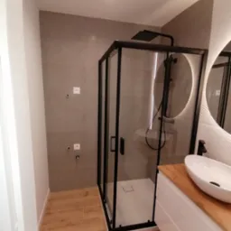 Nowoczesna łazienka po remoncie: kabina prysznicowa z czarną armaturą, umywalka nablatowa na drewnianym blacie, szare płytki ścienne i drewnopodobne płytki podłogowe.