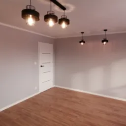 Pomalowane na szaro wnętrze z drewnianą podłogą, oświetlone przez cztery czarne lampy wiszące, w tym jedna podwójna, z białą, minimalistyczną ościeżnicą drzwiową po lewej.