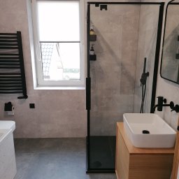 Sebastian Chmiel - Nowoczesna łazienka z szarą płytką, kabiną prysznicową z czarnymi ramami, umywalką nablatową na drewnianej szafce i czarną armaturą.