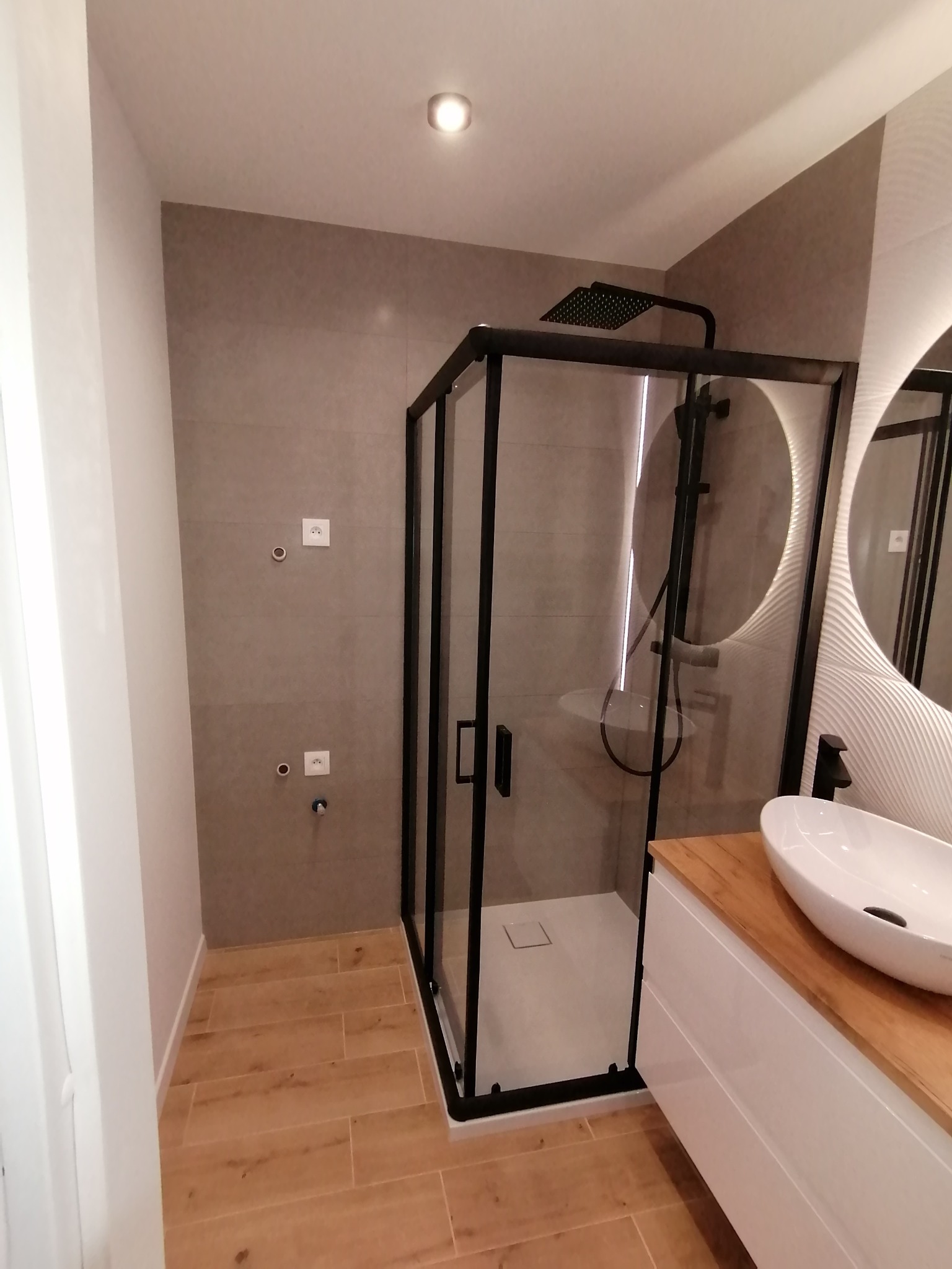 Nowoczesna łazienka po remoncie: kabina prysznicowa z czarną armaturą, umywalka nablatowa na drewnianym blacie, szare płytki ścienne i drewnopodobne płytki podłogowe.