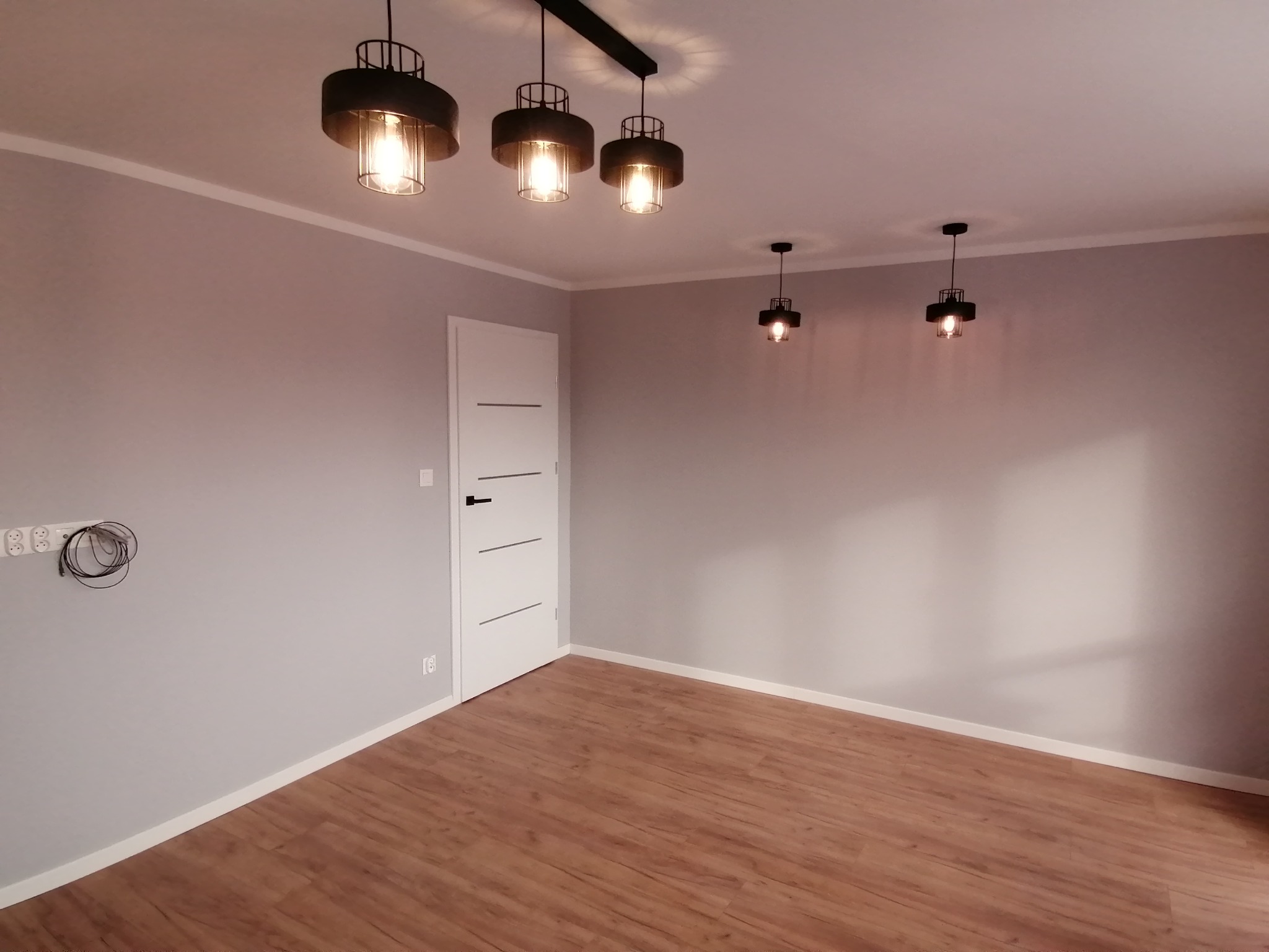 Pomalowane na szaro wnętrze z drewnianą podłogą, oświetlone przez cztery czarne lampy wiszące, w tym jedna podwójna, z białą, minimalistyczną ościeżnicą drzwiową po lewej.