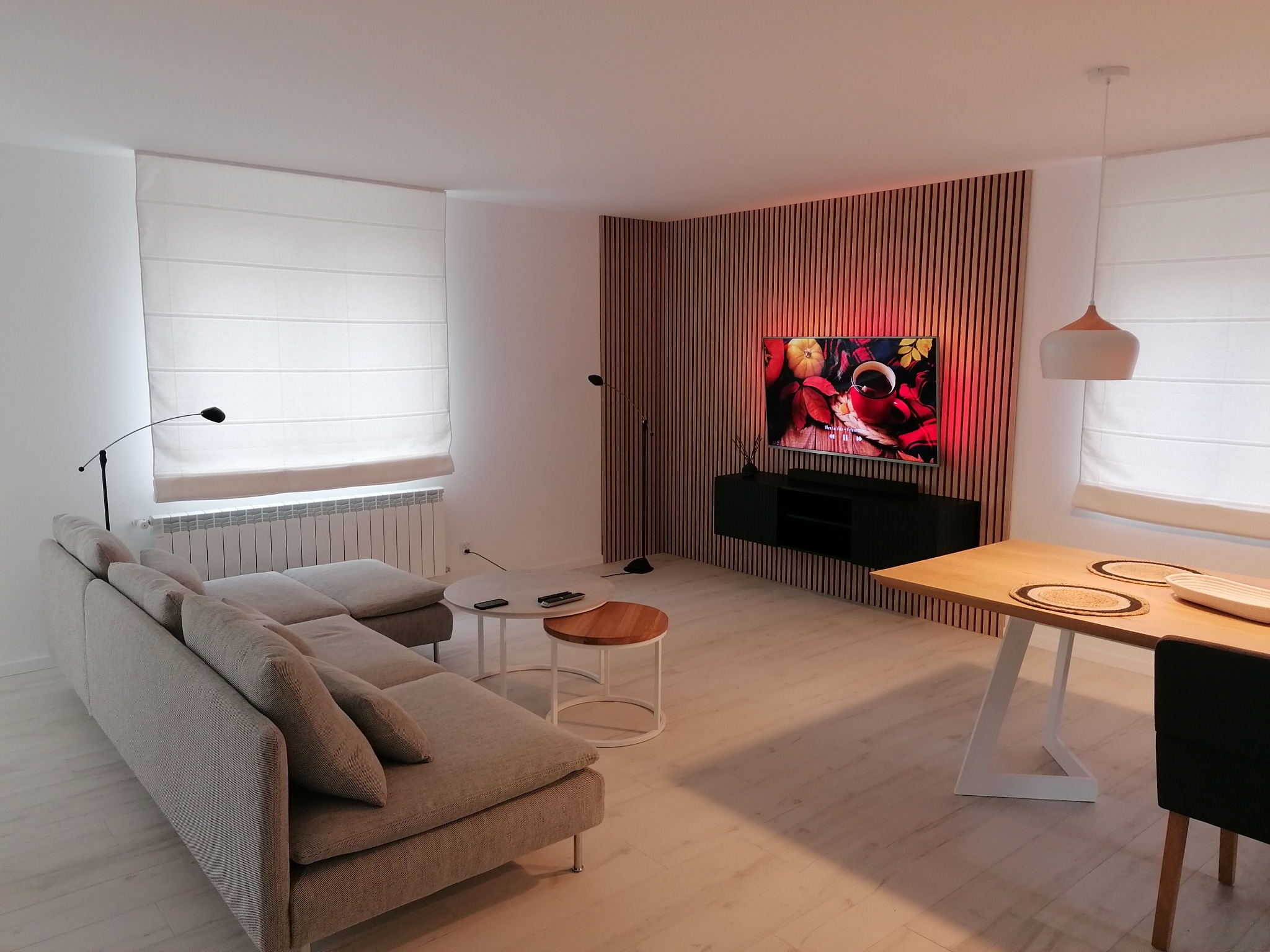 Jasne, przestronne wnętrze salonu z drewnianą ścianą, szarą sofą narożną, okrągłymi stolikami kawowymi i stołem z drewnianym blatem na białej nodze.