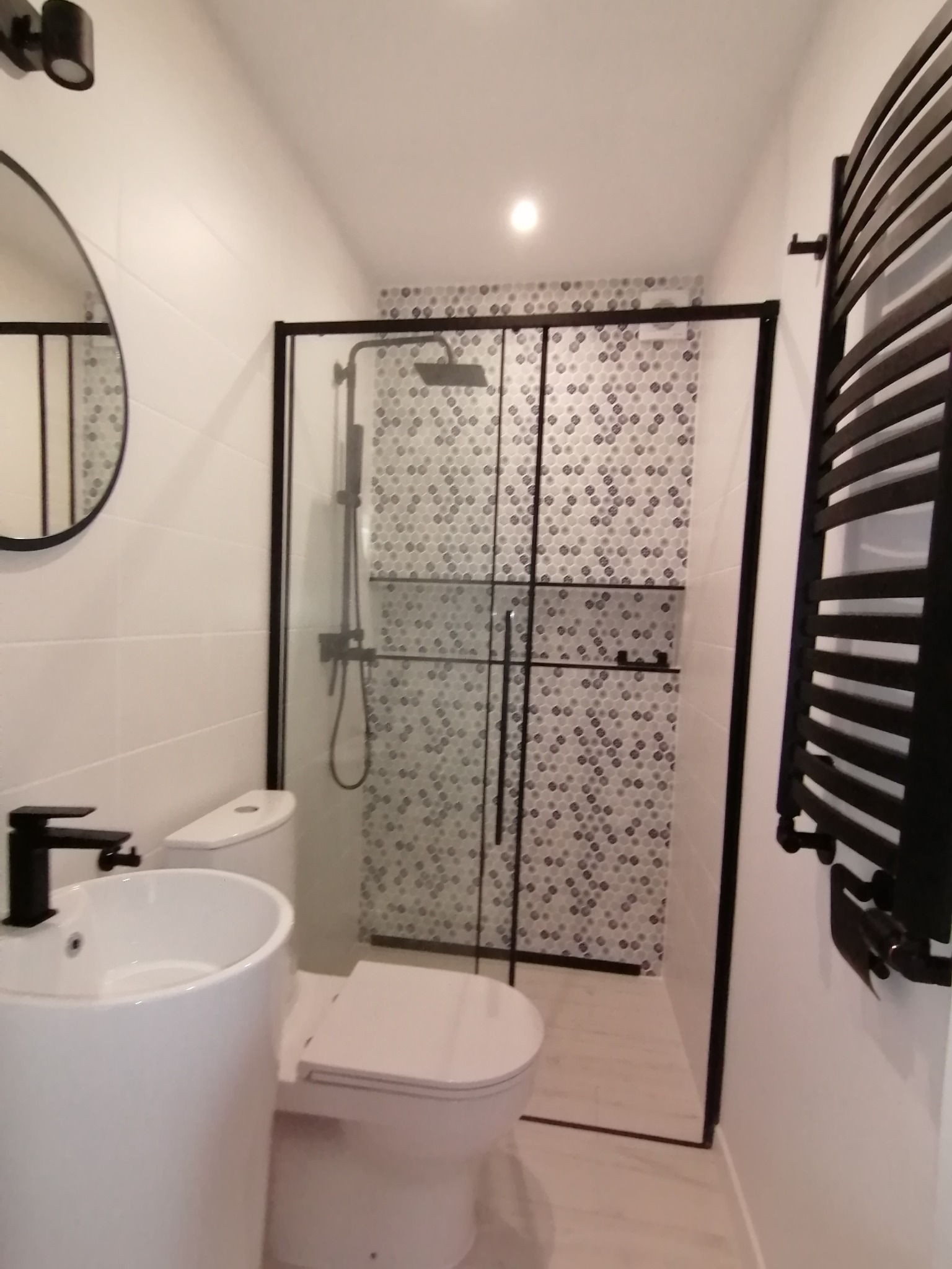 Nowoczesna łazienka z kabiną prysznicową w czarnej ramie, umywalką na postumencie z czarną armaturą, toaletą i grzejnikiem drabinkowym.