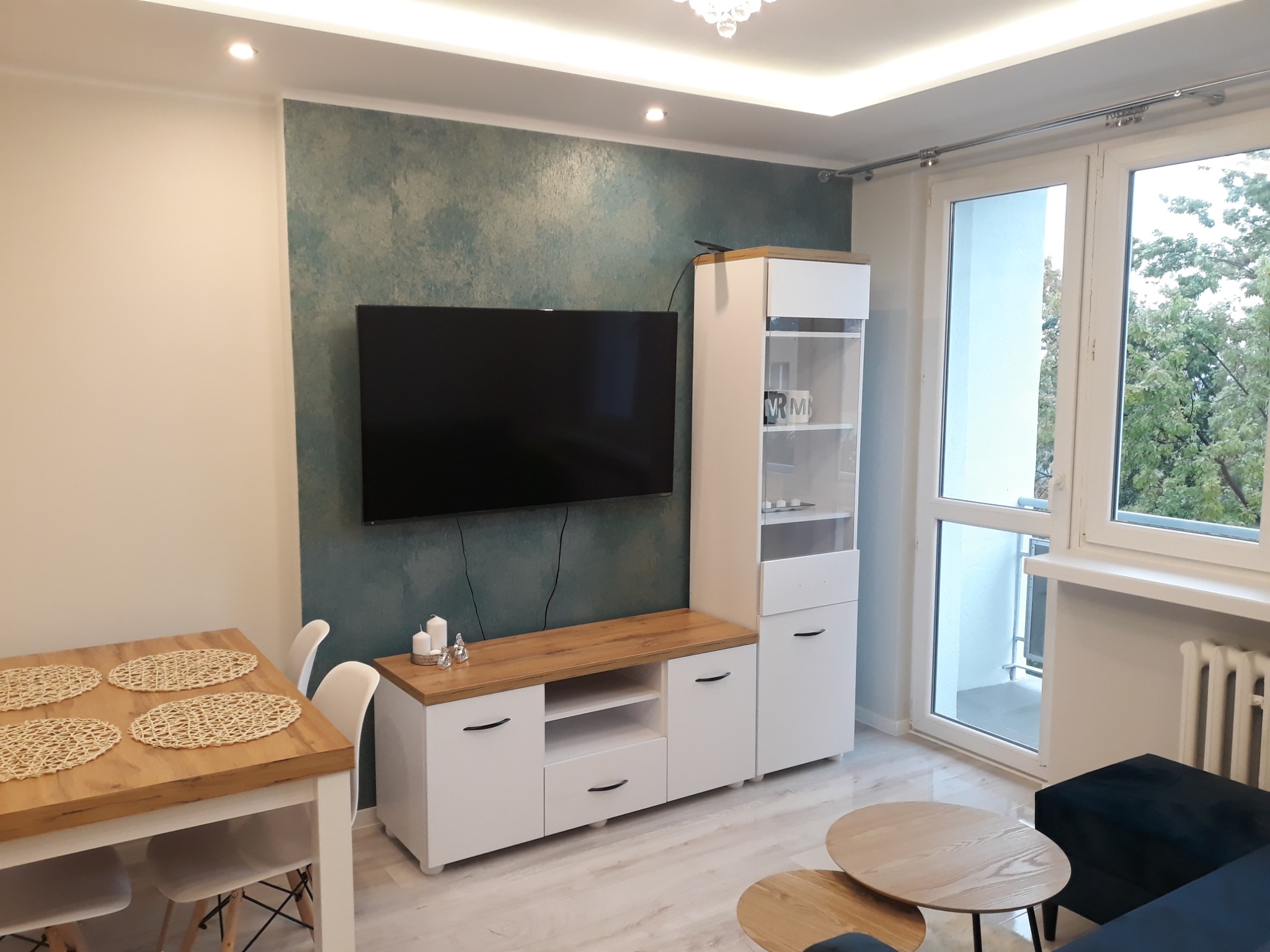 Nowoczesny salon z turkusową ścianą akcentującą, drewnianym stołem z podkładkami, białymi krzesłami i szafką RTV z elementami drewna.