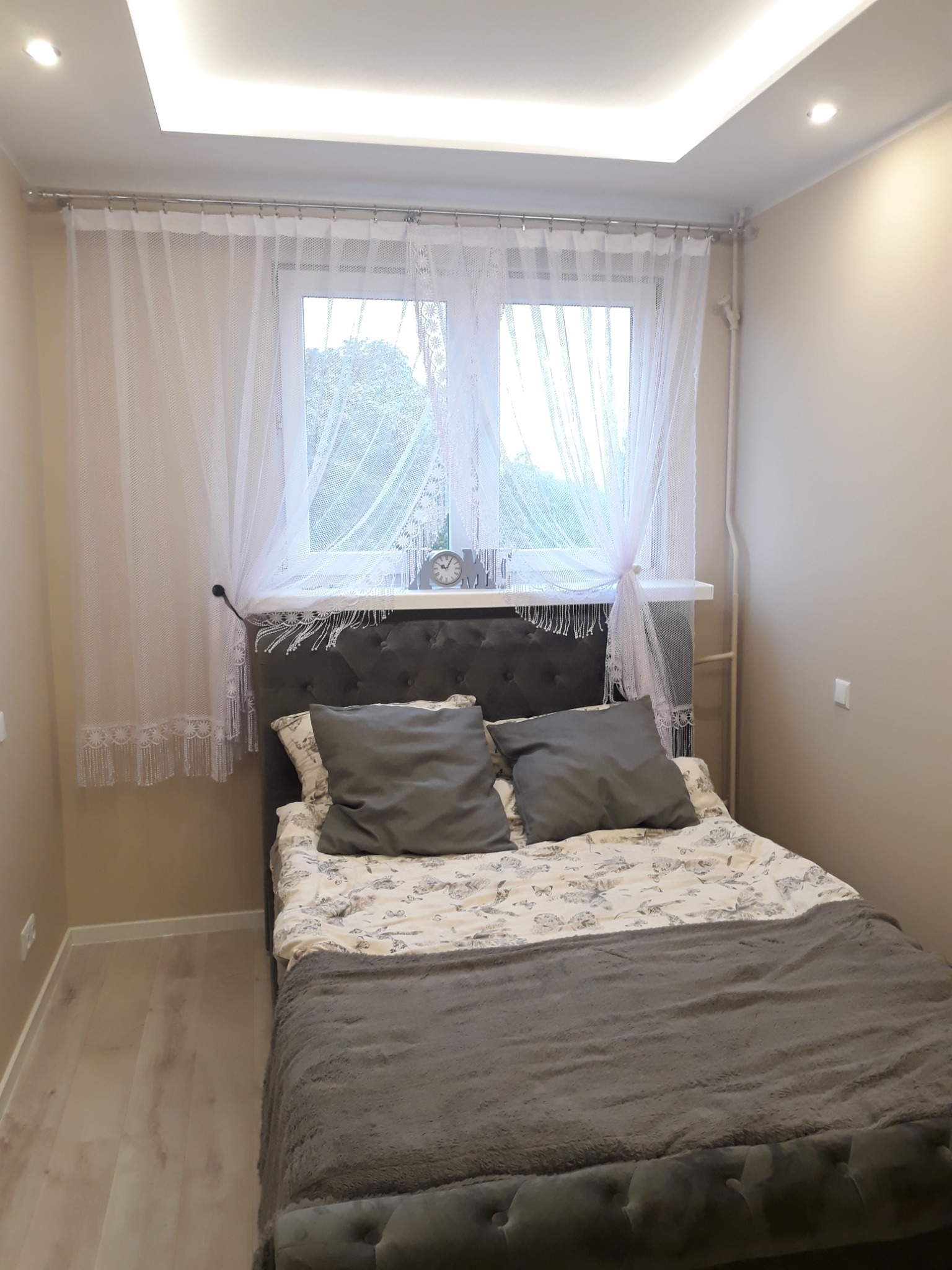 Jasna sypialnia w Sandomierzu po remoncie, z łóżkiem z szarym wezgłowiem, panelami podłogowymi i sufitem podwieszanym z oświetleniem LED.