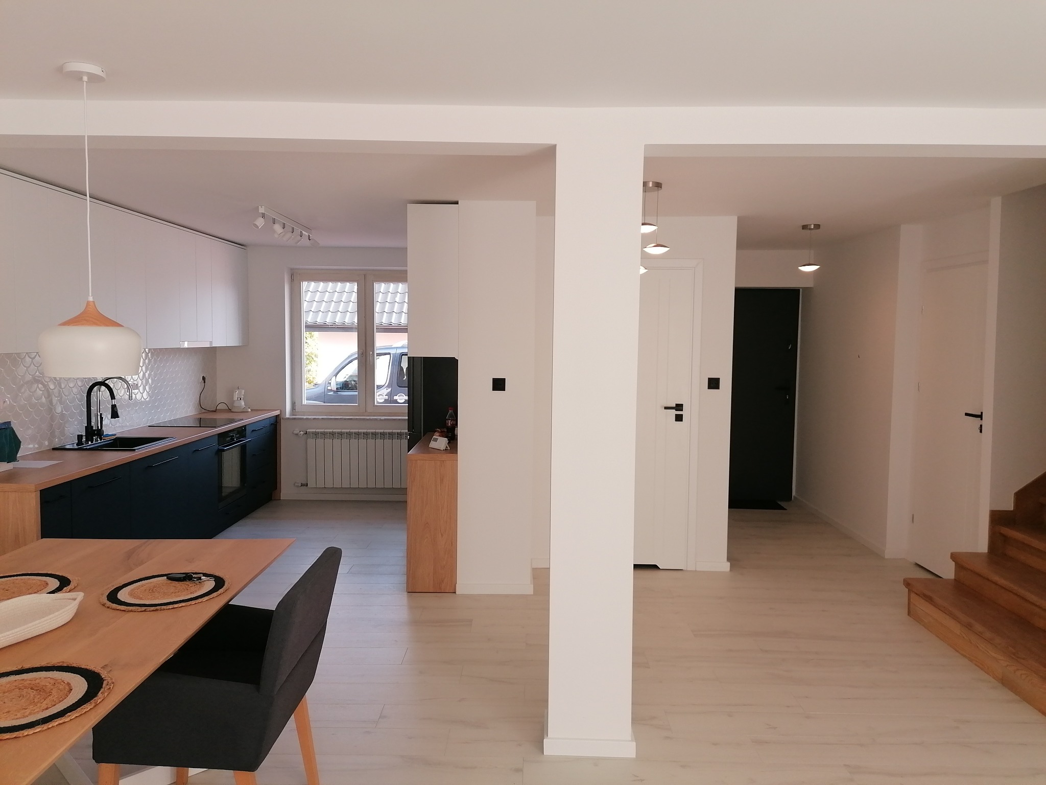 Nowoczesne wnętrze domu z otwartą kuchnią, drewnianym stołem i schodami, utrzymane w jasnej kolorystyce z akcentami granatu i drewna.