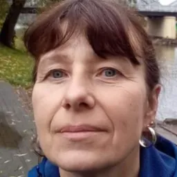 Portret kobiety z krótkimi brązowymi włosami, srebrnymi kolczykami i niebieską bluzą na tle rzeki i mostu.