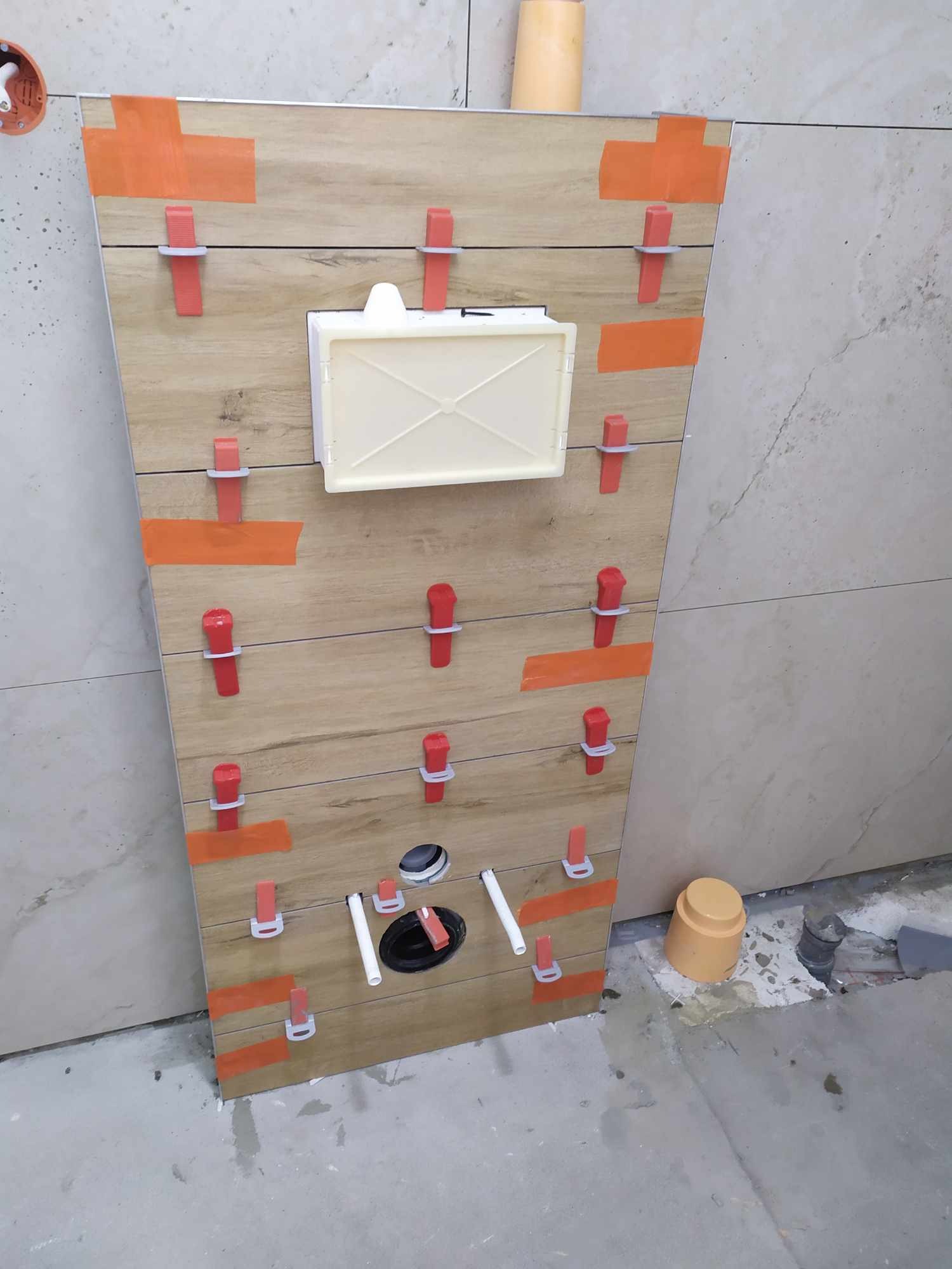 Montaż płytek imitujących drewno na ścianie z zamontowaną podtynkową spłuczką, użycie klipsów poziomujących i pomarańczowej taśmy maskującej, widoczne rury hydrauliczne.