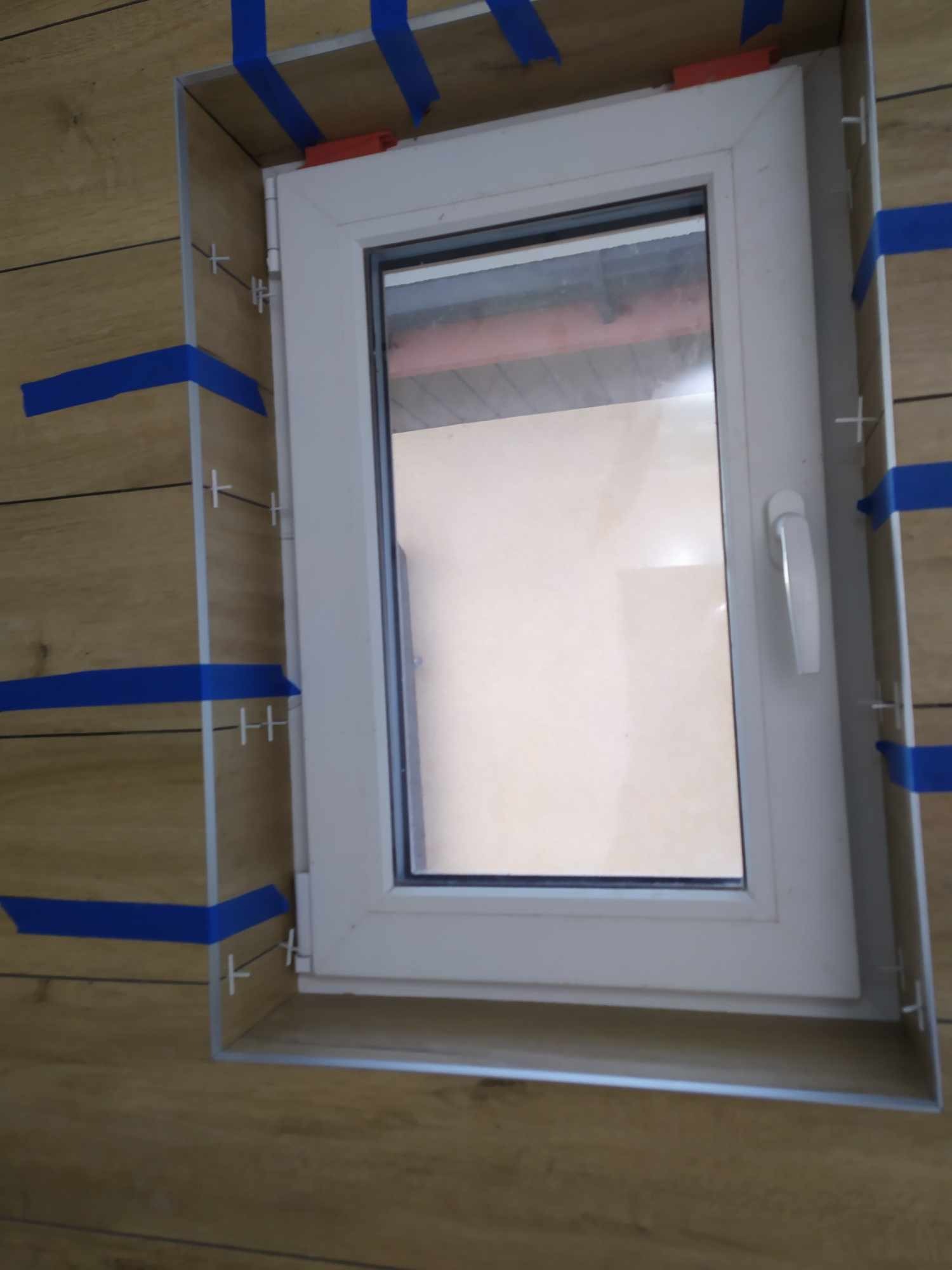 Montaż płytek drewnopodobnych wokół okna, użycie taśmy malarskiej i krzyżyków dystansowych, widoczny biały profil okienny.