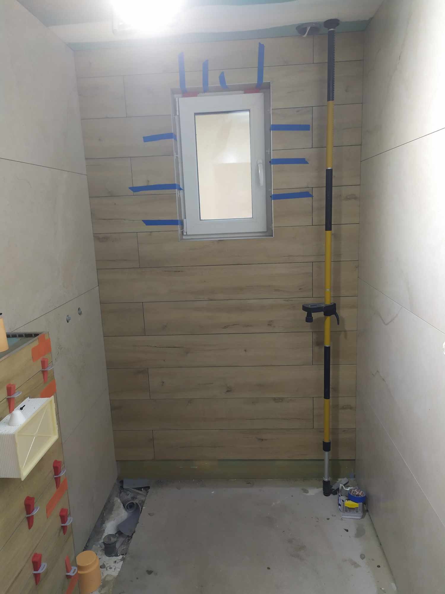 Wnętrze łazienki w trakcie prac wykończeniowych: ściany wyłożone płytkami imitującymi drewno, okno zabezpieczone taśmą malarską, widoczny poziomujący laser na tyczce oraz system poziomowania płytek...