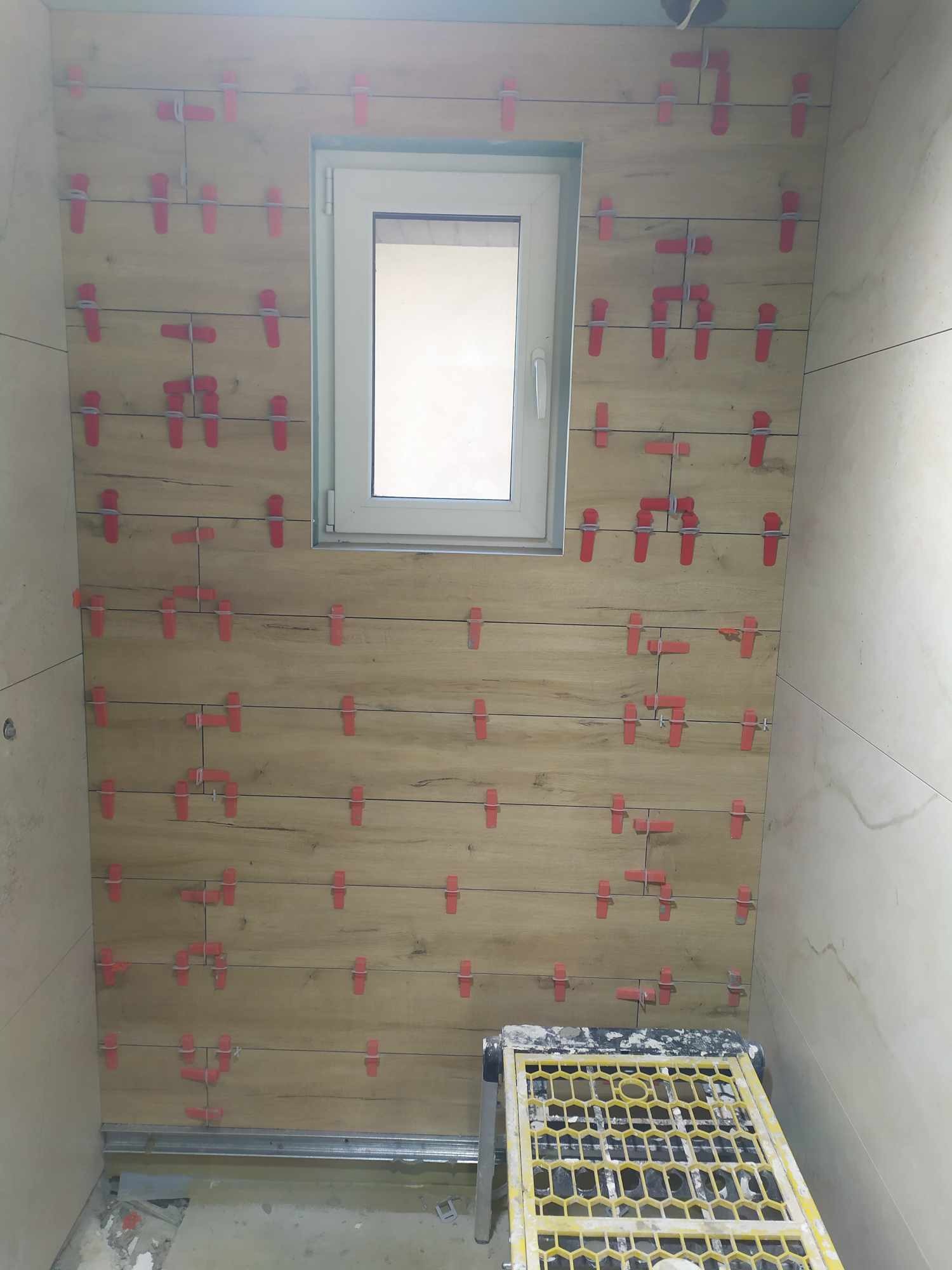 Ściana w trakcie układania płytek imitujących drewno, z widocznymi czerwonymi klipsami poziomującymi, małe okno w tle, żółty podest roboczy na dole kadru.