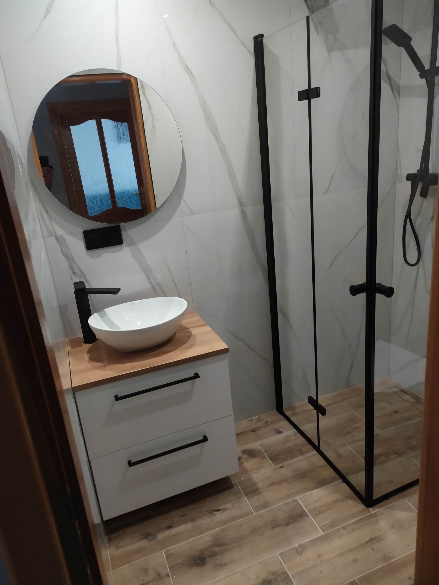 Nowoczesna łazienka z kabiną prysznicową w czarnej ramie, okrągłym lustrem nad białą szafką z drewnianym blatem i umywalką nablatową, marmurowe płytki na ścianach oraz podłoga imitująca drewno.