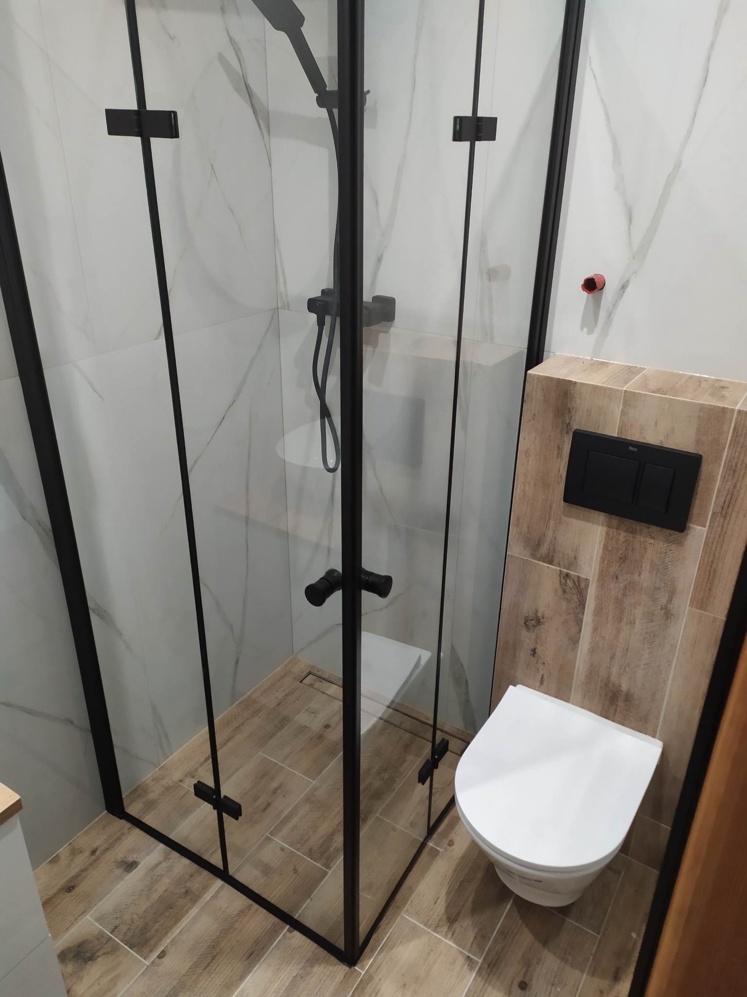 Nowoczesna łazienka z kabiną prysznicową z czarnymi profilami, podwieszaną toaletą i płytkami imitującymi drewno na podłodze oraz marmur na ścianach.