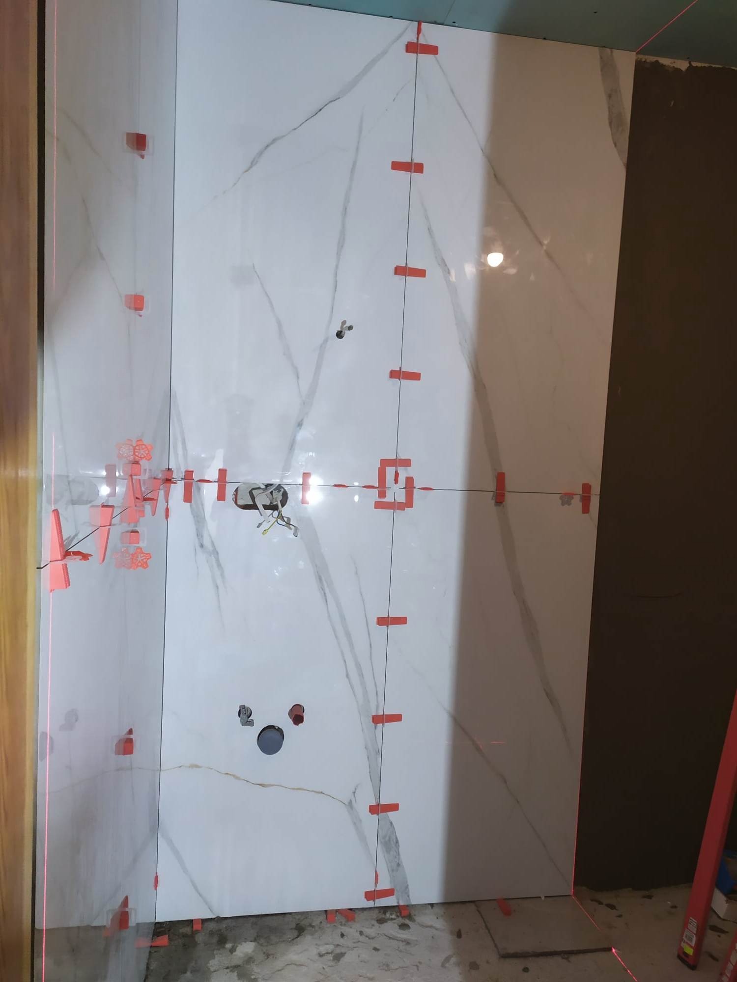 Ściana w trakcie układania wielkoformatowych płytek imitujących marmur, z widocznymi plastikowymi krzyżykami dystansowymi i laserowym poziomem wyznaczającym linię, otwory pod instalacje.