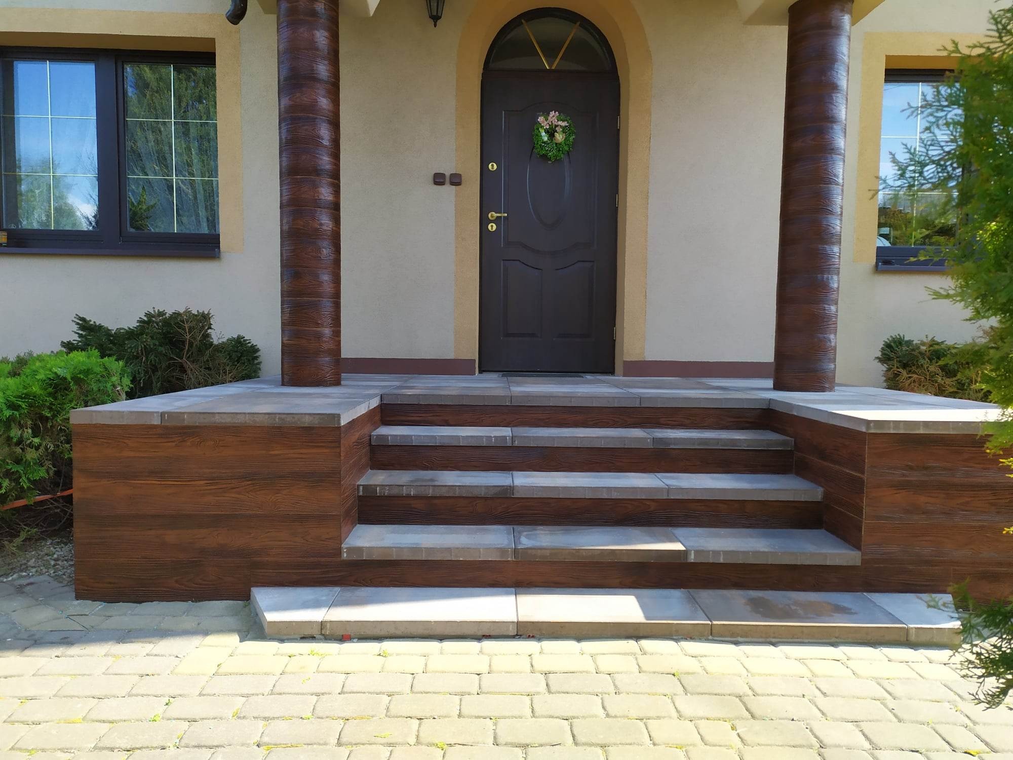 Wejście do domu z brązowymi drzwiami, zdobionymi wieńcem, oraz schodami wykończonymi płytkami imitującymi drewno i szarym kamieniem, z kolumnami o podobnym wykończeniu.