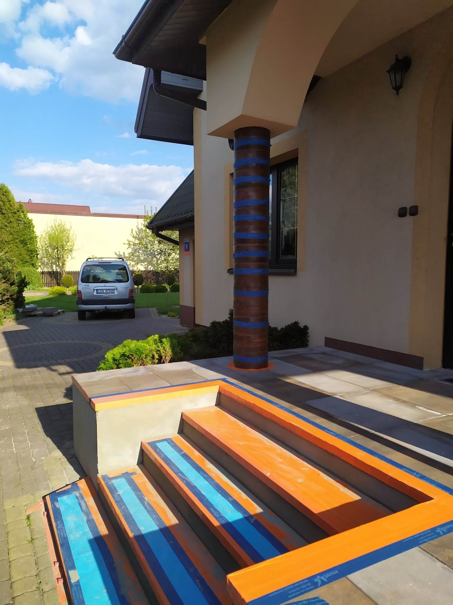 Schody wejściowe do domu w trakcie malowania, zabezpieczone niebieską taśmą malarską, kolumna wejściowa z imitacją drewna również z taśmą, na podjeździe zaparkowany srebrny samochód.