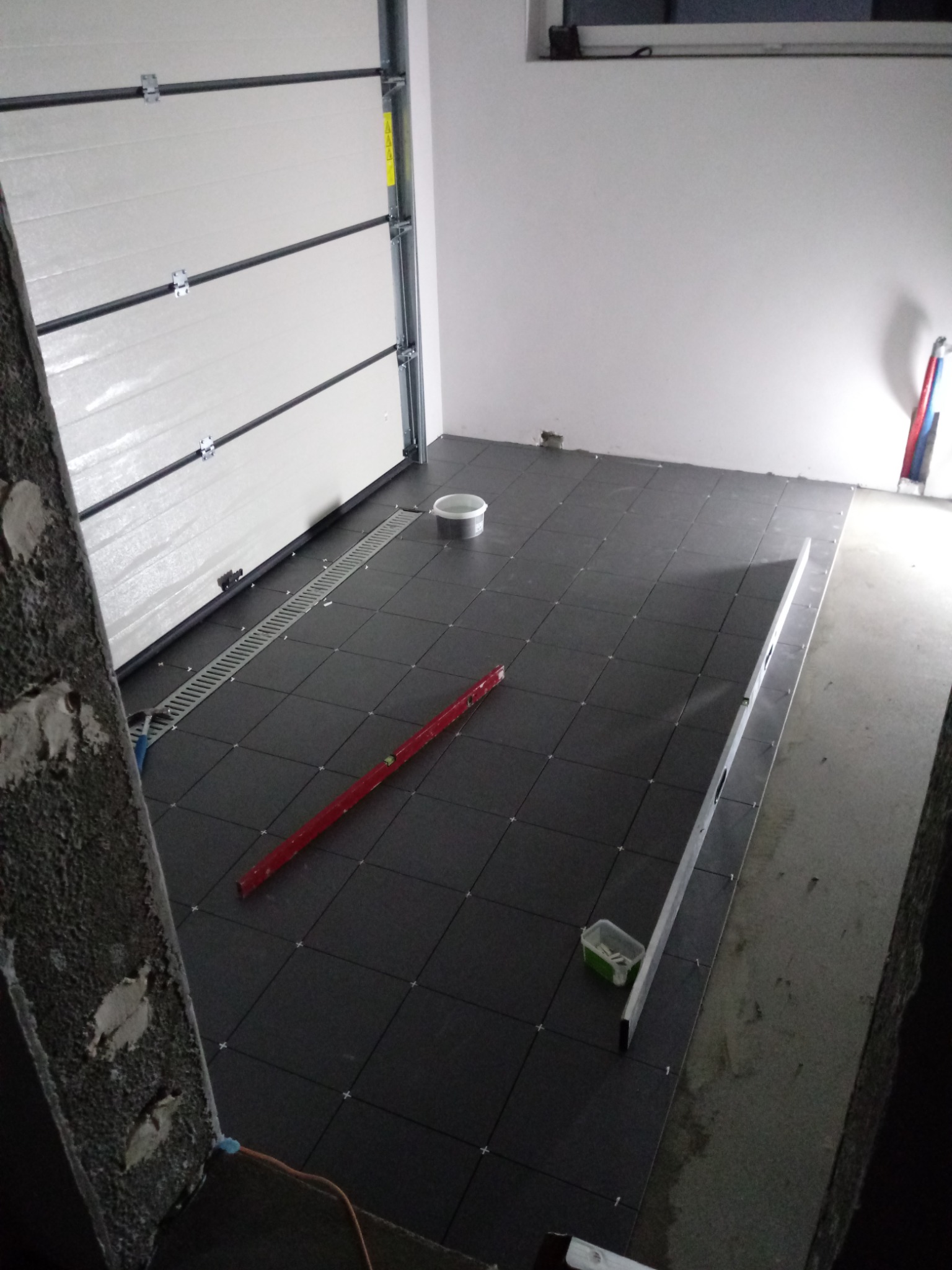 Układanie ciemnych płytek podłogowych w garażu, widoczna kratka odpływowa, poziomica czerwona i srebrna, krzyżyki dystansowe, surowy beton na ścianach, brama garażowa segmentowa.