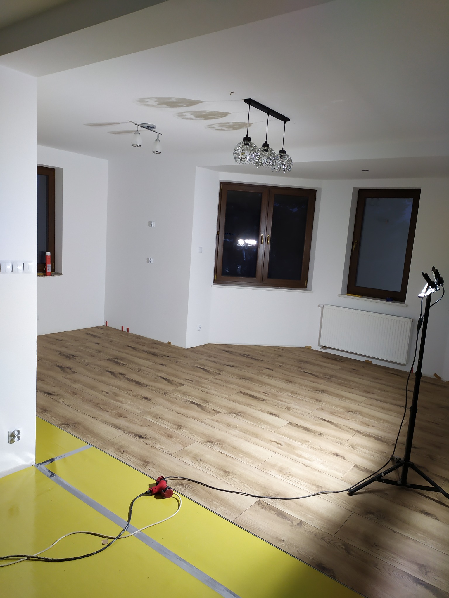 Układanie paneli podłogowych w jasnym wnętrzu w Kąkolewnicy, widoczna folia podkładowa, lampa budowlana i narzędzia.