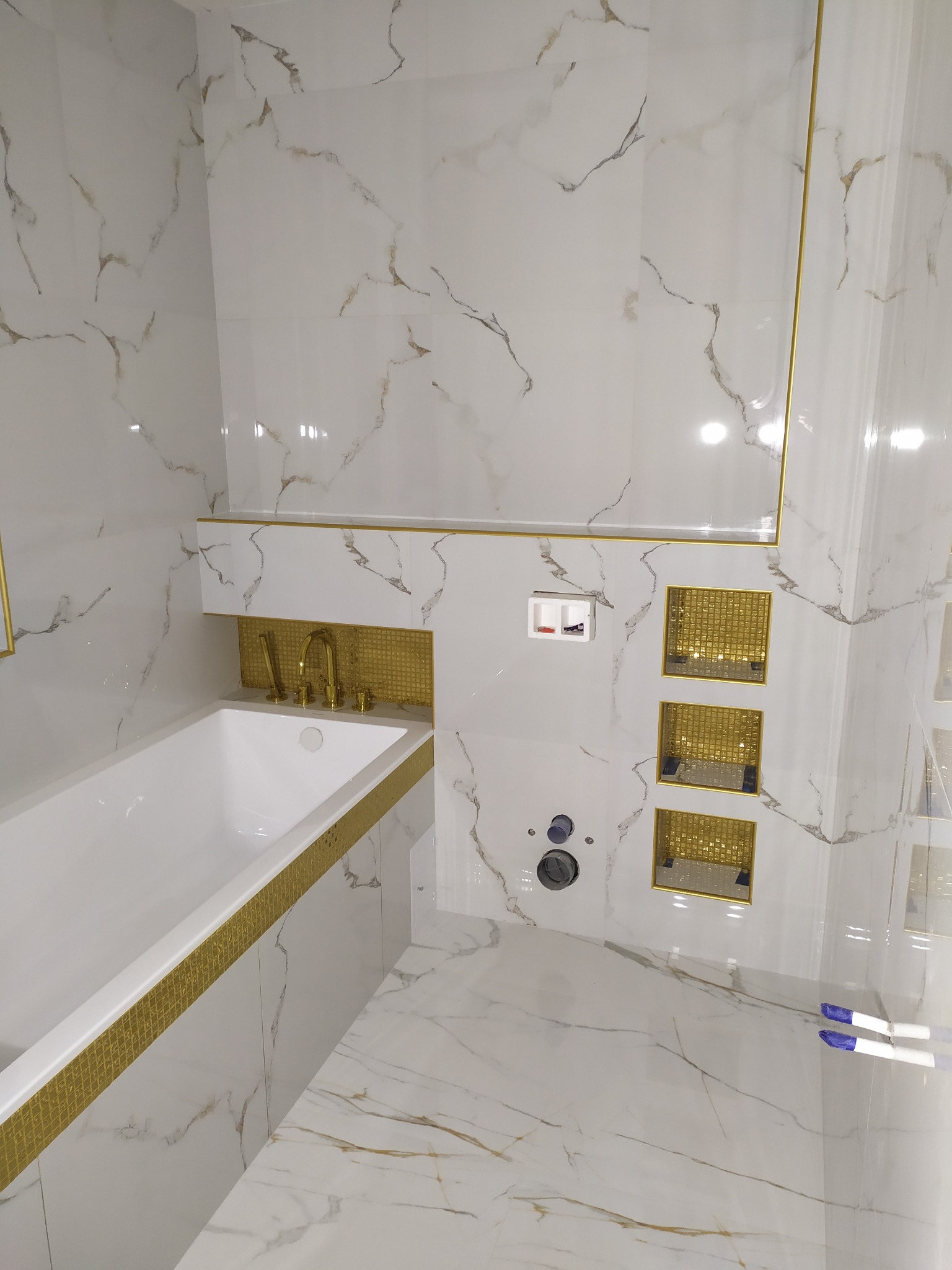Wykończona łazienka z białymi płytkami imitującymi marmur z złotymi żyłkami, złotymi wstawkami mozaikowymi wokół wanny i wnęk ściennych, widoczne elementy instalacji wodno-kanalizacyjnej w trakcie...