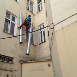 IgloWent - Pracownik na linach montuje metalowe rury wentylacyjne na elewacji budynku z oknami i starą klimatyzacją.