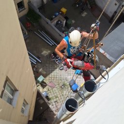 IgloWent - Pracownik w uprzęży alpinistycznej, z widoku z góry, instaluje rury wentylacyjne na zewnętrznej ścianie budynku mieszkalnego w podwórzu.