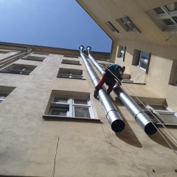 IgloWent - Pracownik na linie alpinistycznej montuje dwie rury wentylacyjne na elewacji budynku mieszkalnego, widok z dołu, niebieskie niebo w tle.