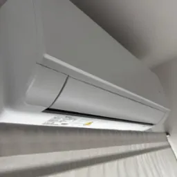 Biały klimatyzator ścienny, widoczny od dołu, zamontowany w pomieszczeniu z zasłonami. Widoczne naklejki informacyjne na urządzeniu.