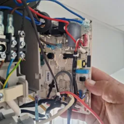 Serwisant trzyma płytę sterującą klimatyzatora z widocznymi podłączeniami elektrycznymi i elementami elektronicznymi, na tle sufitu i okna.