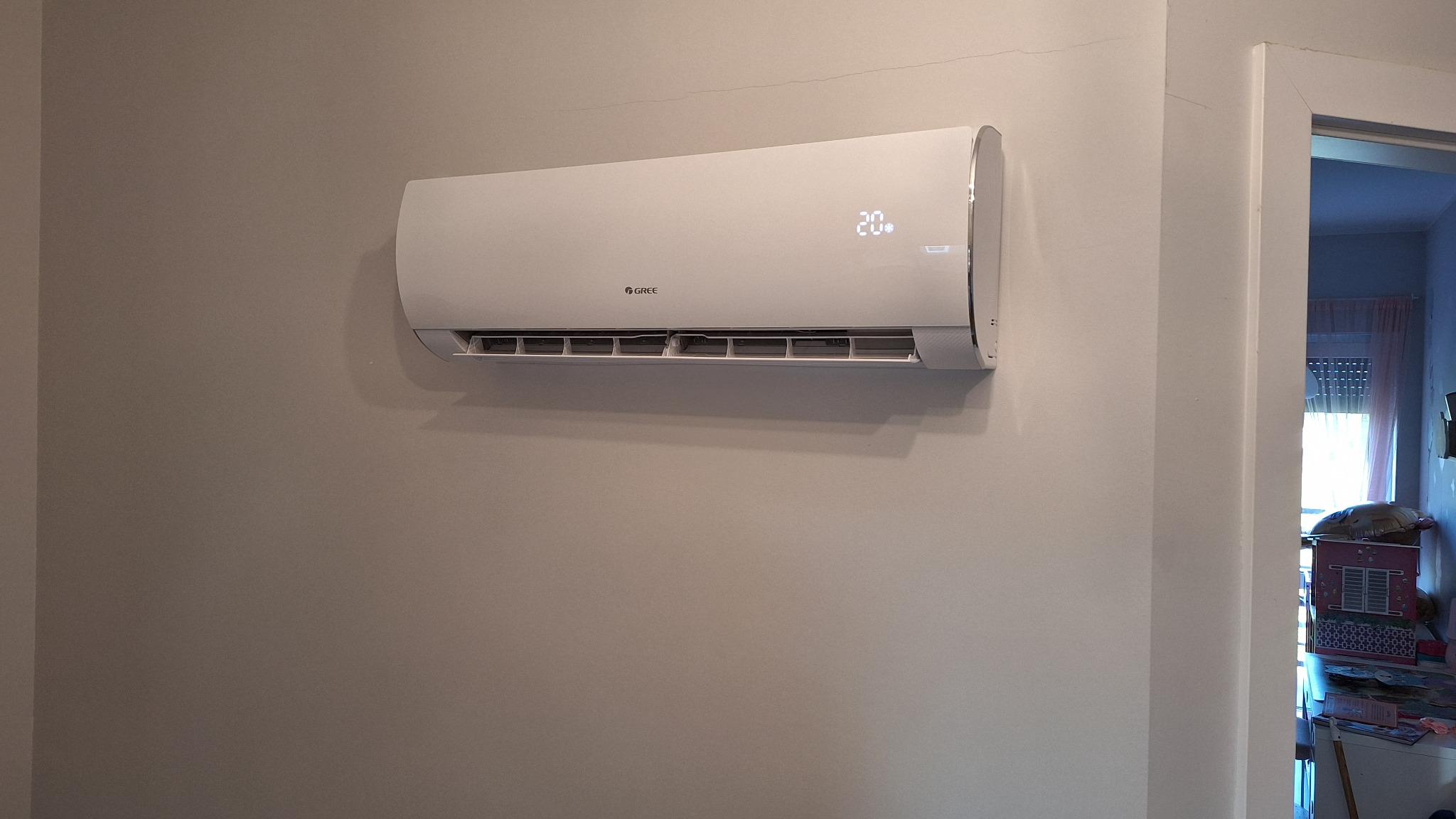 Biały klimatyzator marki Gree zamontowany na szarej ścianie w pokoju, wyświetlający temperaturę 20 stopni Celsjusza, widoczne pęknięcie na ścianie powyżej klimatyzatora.