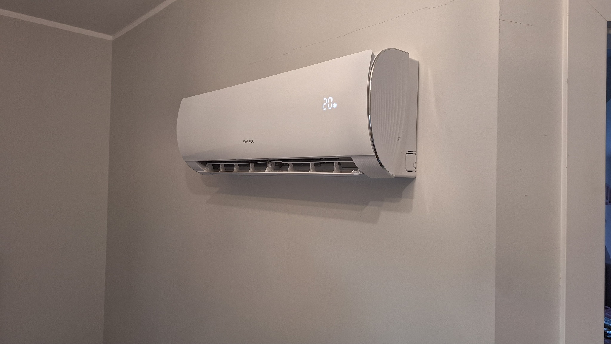 Biały klimatyzator marki Gree z wyświetlaczem wskazującym temperaturę 20 stopni, zamontowany na szarej ścianie w rogu pomieszczenia, widoczne pęknięcie na ścianie przy suficie.