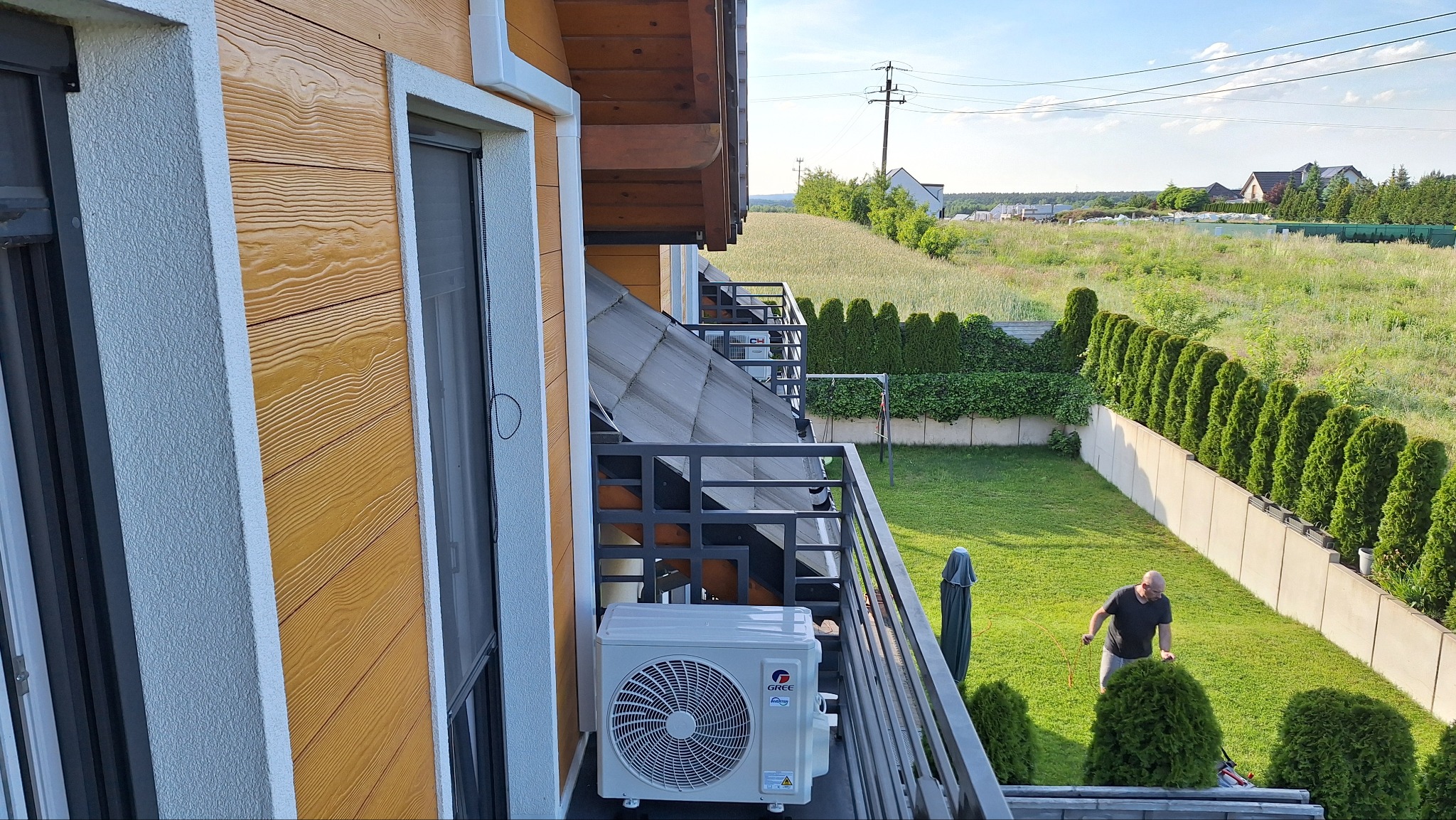 Biała jednostka zewnętrzna klimatyzatora Gree zamontowana na balkonie domu z drewnianą elewacją, widok na zadbany trawnik i mężczyznę z kosiarką.