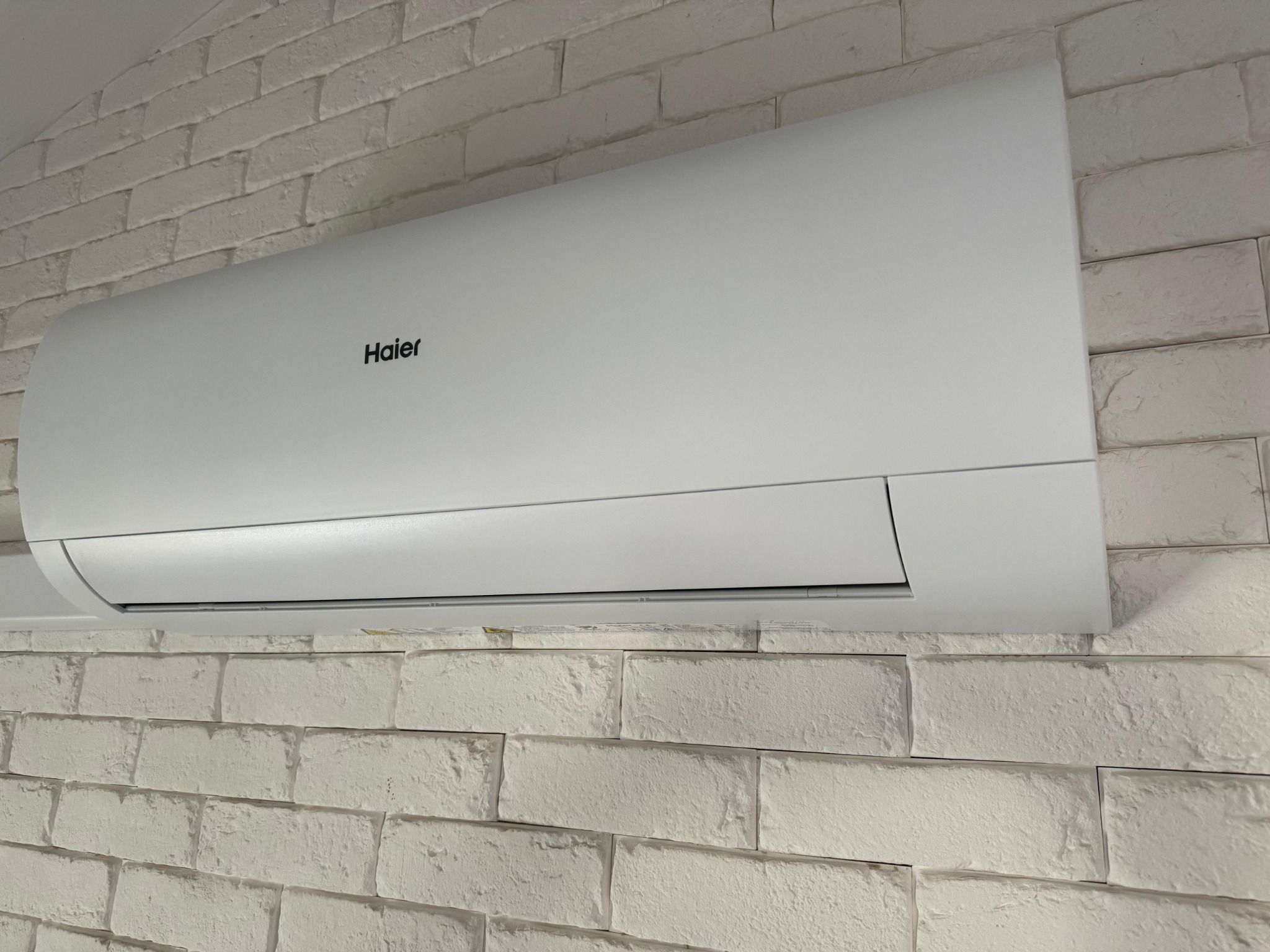 Biała jednostka wewnętrzna klimatyzatora Haier zamontowana na ścianie z białej cegły dekoracyjnej.