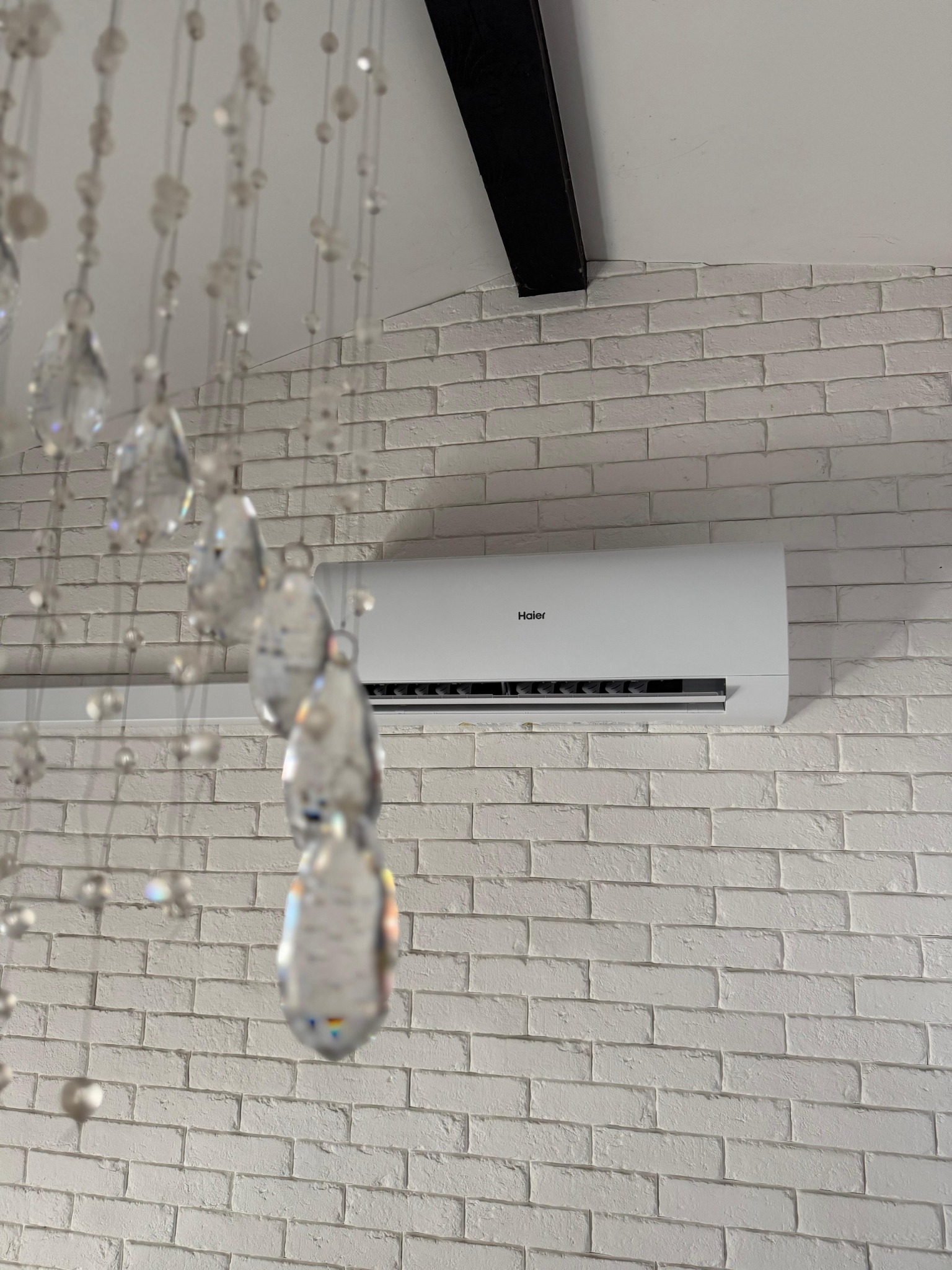 Biały klimatyzator Haier zamontowany na ścianie z białej cegły, widok z dołu, kryształowy żyrandol w pierwszym planie.