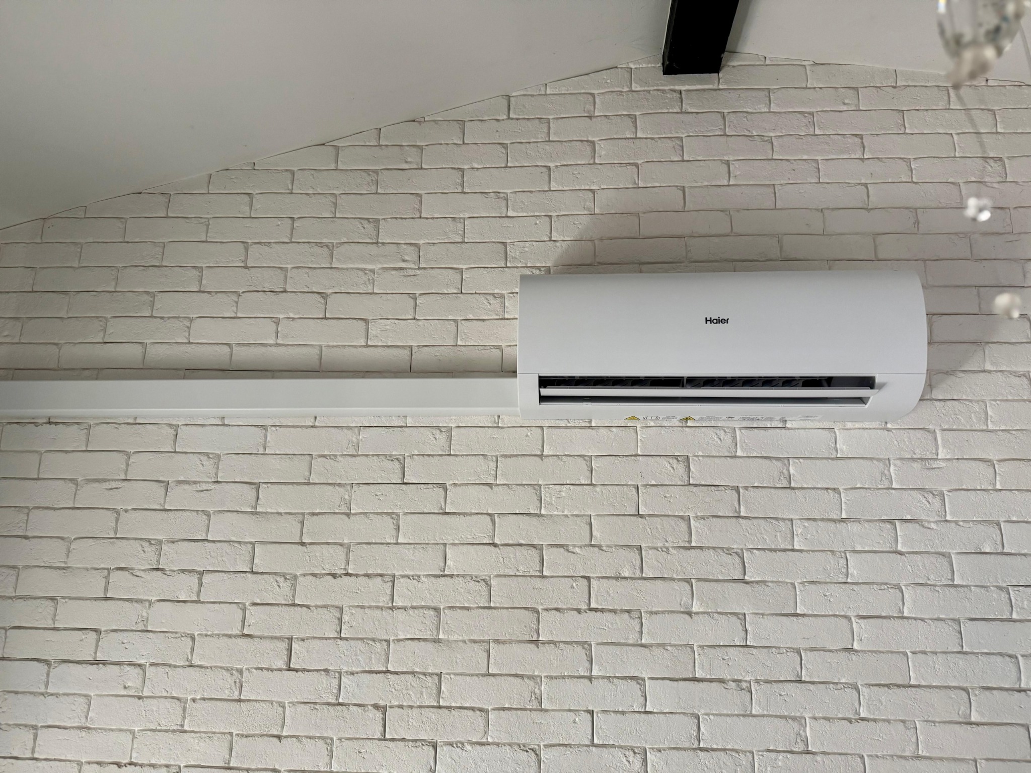 Biały klimatyzator Haier zamontowany na ścianie z białej cegły w pomieszczeniu ze skośnym sufitem.