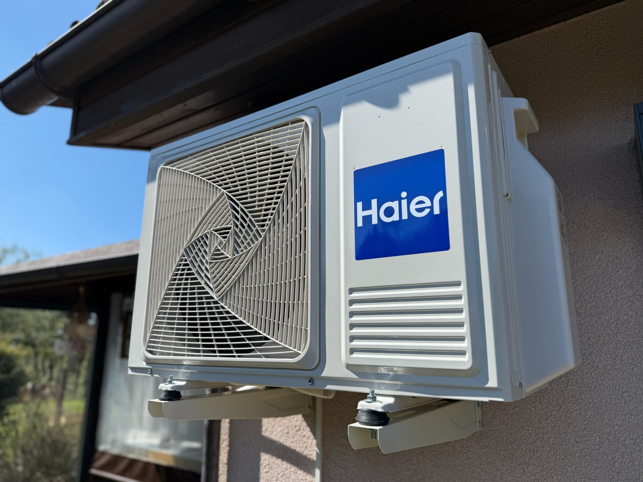 Zewnętrzna jednostka klimatyzacji Haier zamontowana na ścianie budynku z widocznymi wspornikami i gumowymi podkładkami antywibracyjnymi, na tle zieleni i błękitnego nieba.