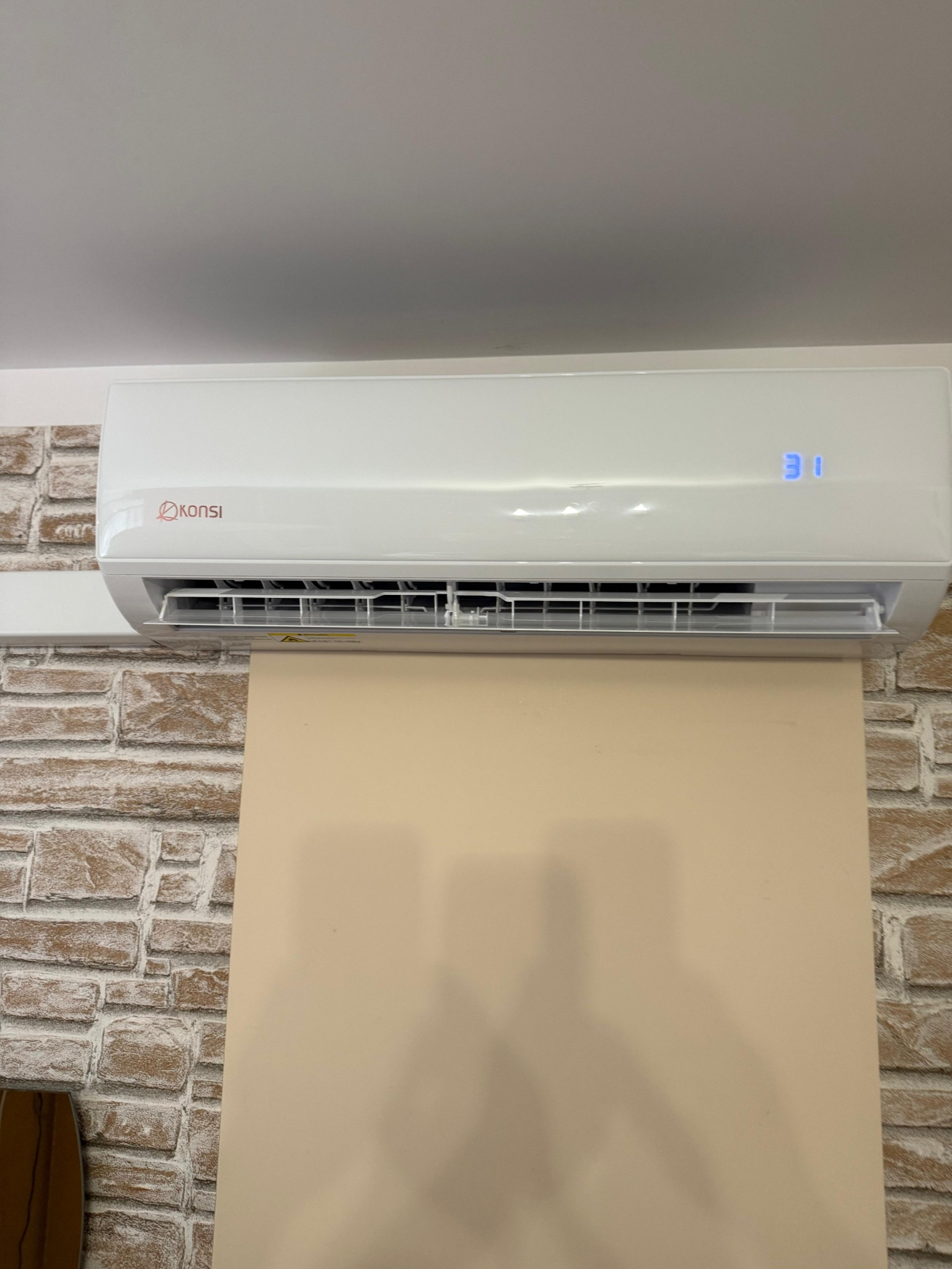 Biały klimatyzator marki Konsi zamontowany na ścianie z dekoracyjną cegłą, wyświetlający temperaturę 31 stopni.