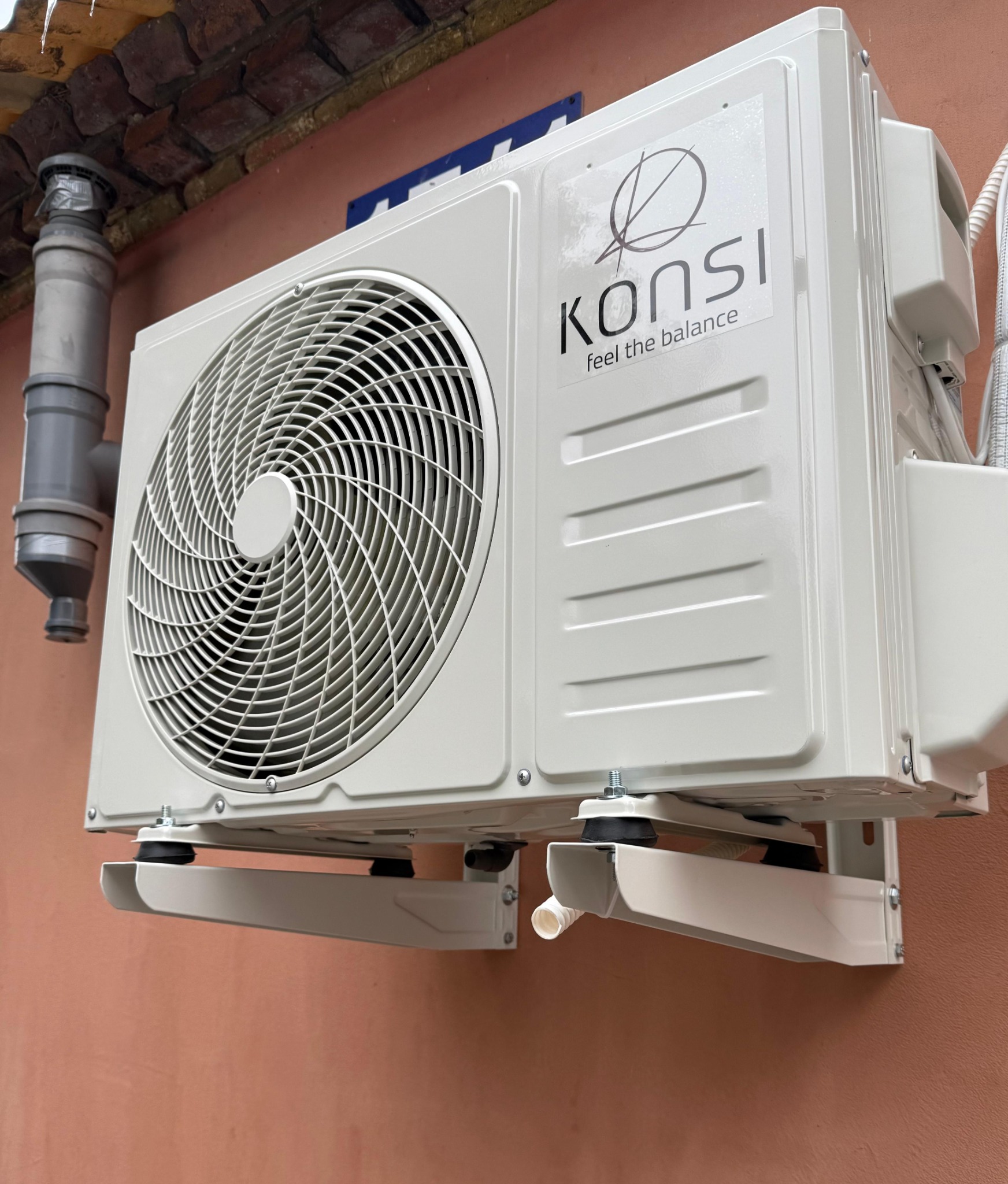 Biała jednostka zewnętrzna klimatyzacji marki KONSI zamontowana na ścianie budynku z brązową elewacją, widoczny odpływ skroplin oraz fragment instalacji wentylacyjnej.