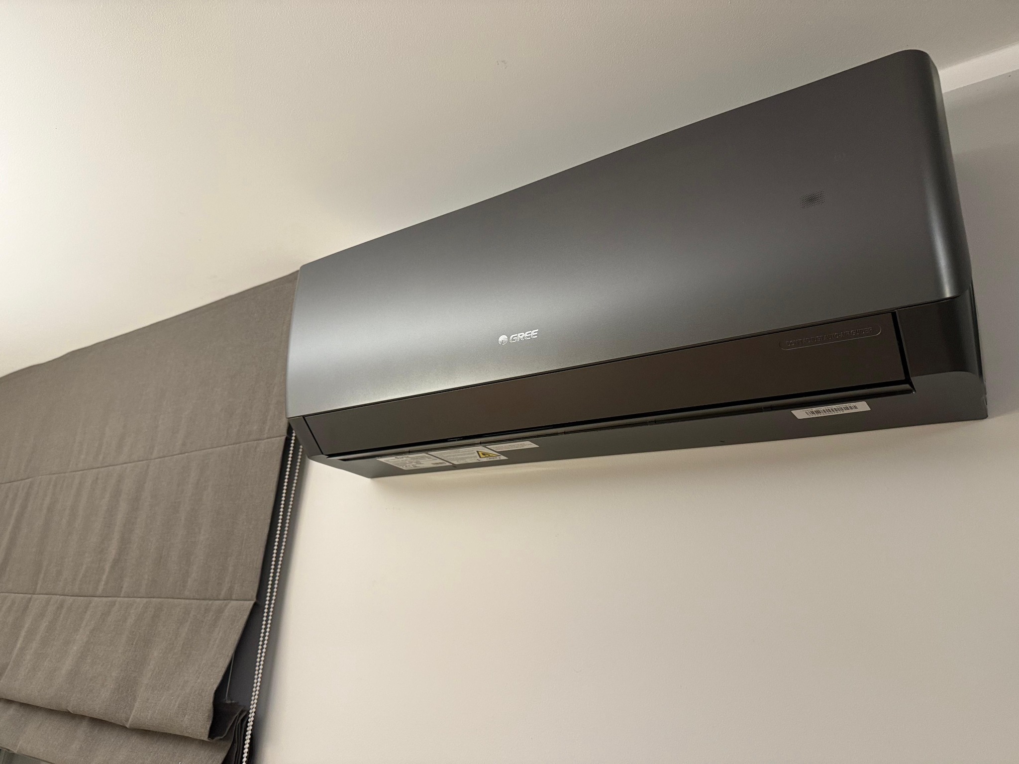 Grafitowa jednostka klimatyzacyjna Gree zamontowana na białej ścianie, obok zasłony rzymskiej w odcieniach szarości.