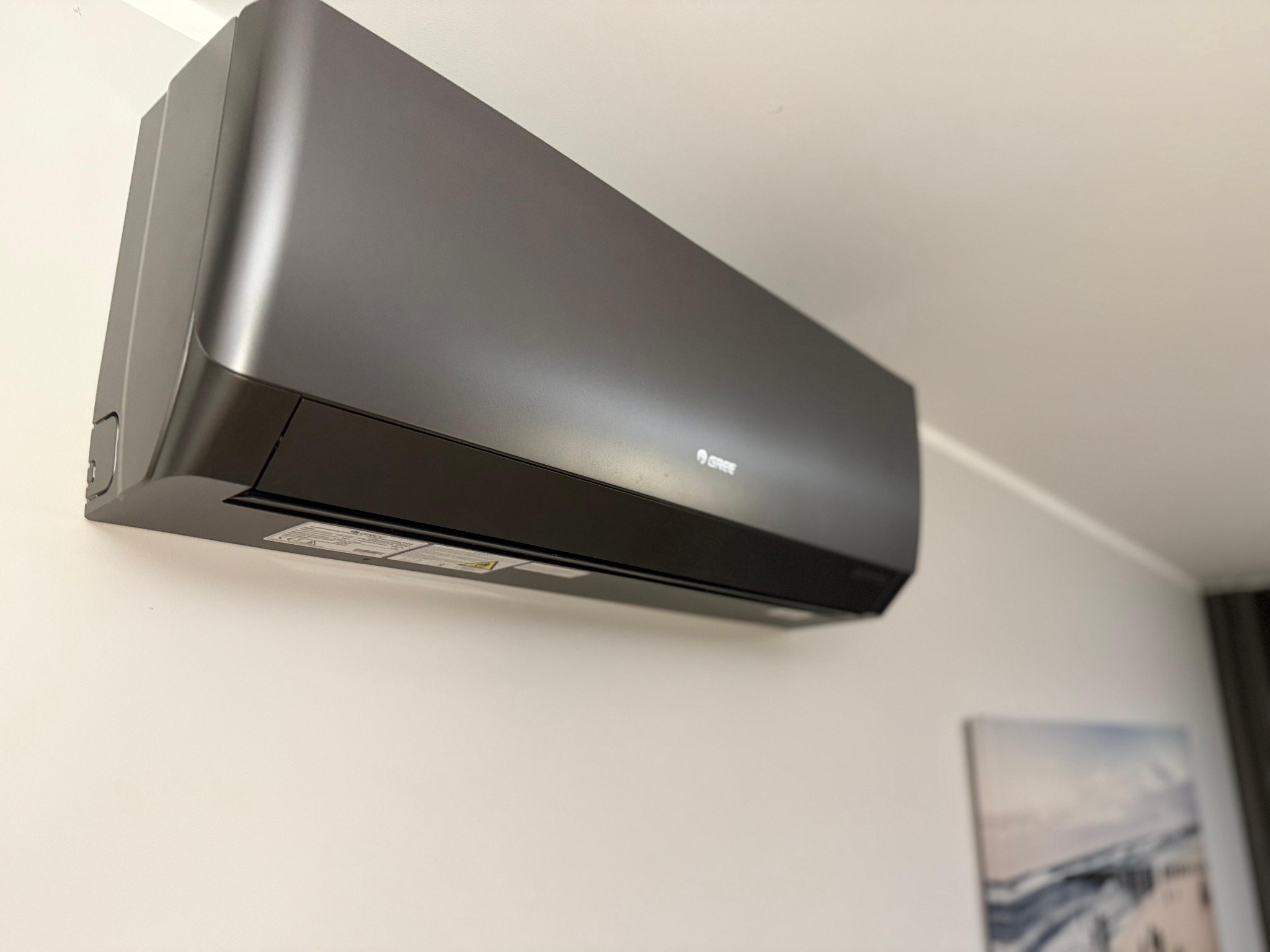 Grafitowy klimatyzator marki Gree zamontowany na białej ścianie w pomieszczeniu z obrazem w tle.
